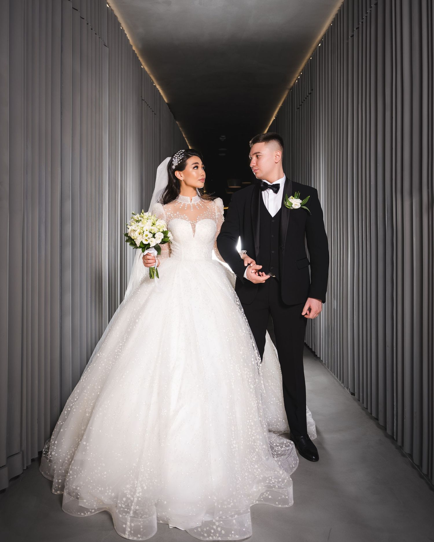 Bride in white ball gown with veil holding bouquet walks with groom in black tuxedo down modern corridor.