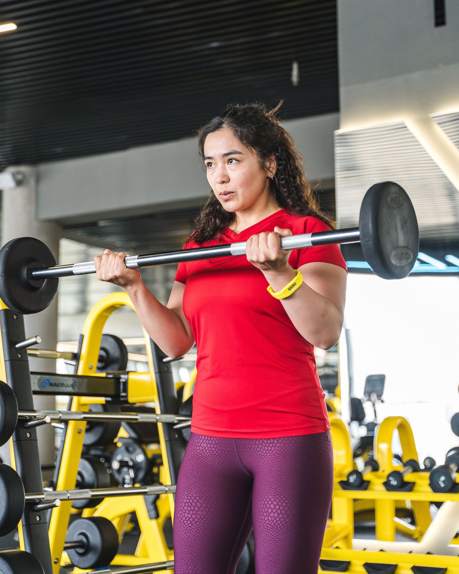 Person in red top doing barbell squats during workout session in modern fitness facility.