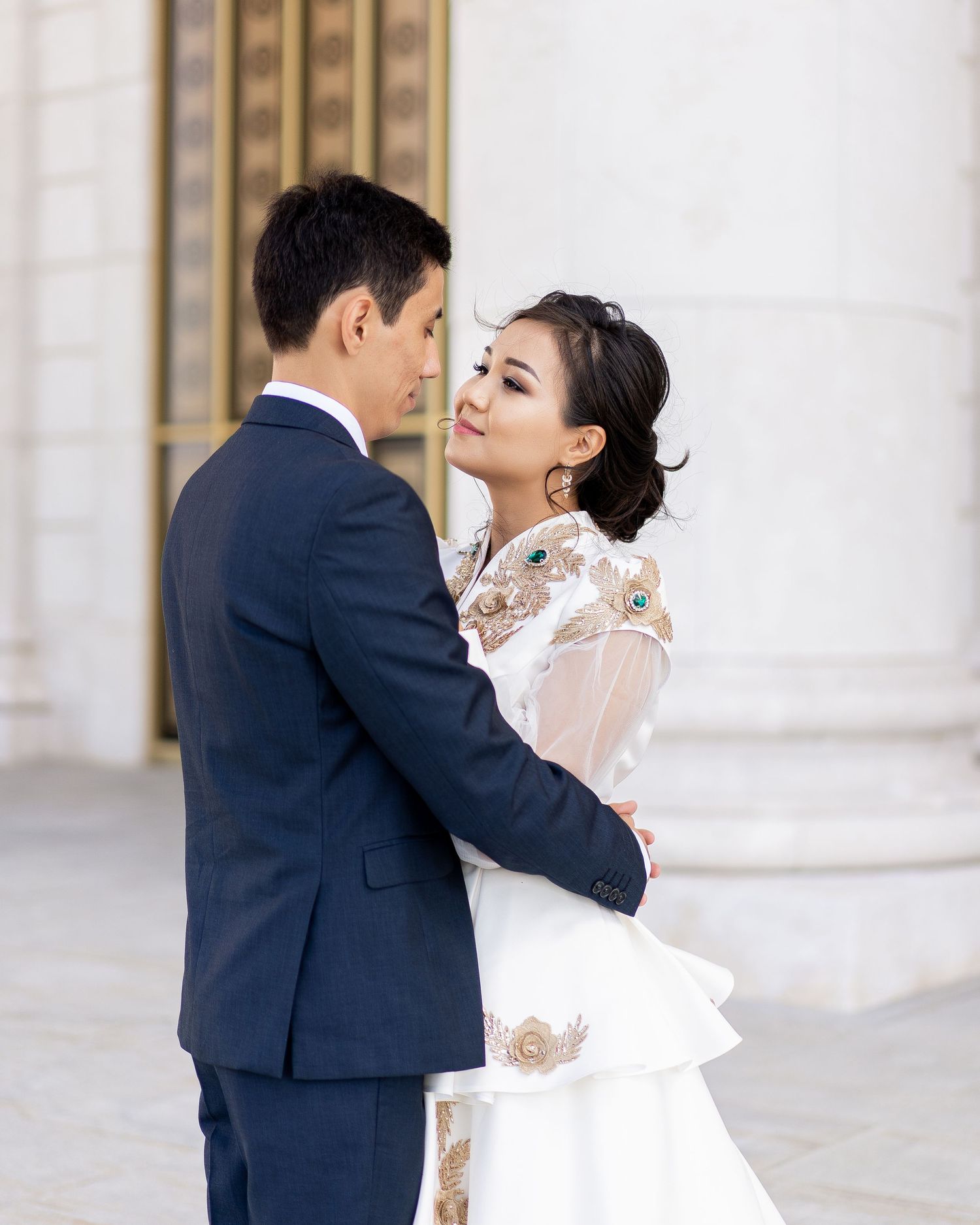 A couple in wedding attire embraces outdoors, the bride in a white lace gown looking over her shoulder.