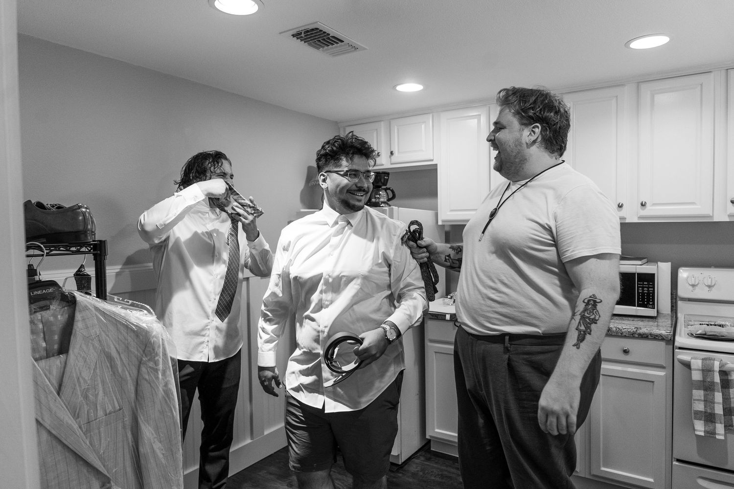 Three men in dress shirts socializing in a kitchen, one smiling and holding a belt in a black and white photo.