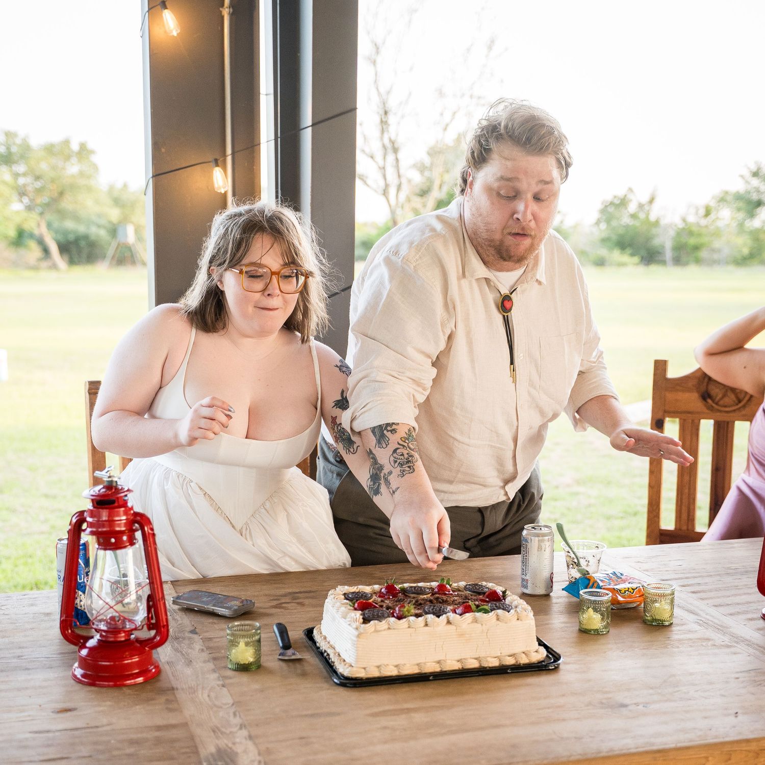 A tattooed groom and bride in white cut their wedding cake at a rustic outdoor reception table.