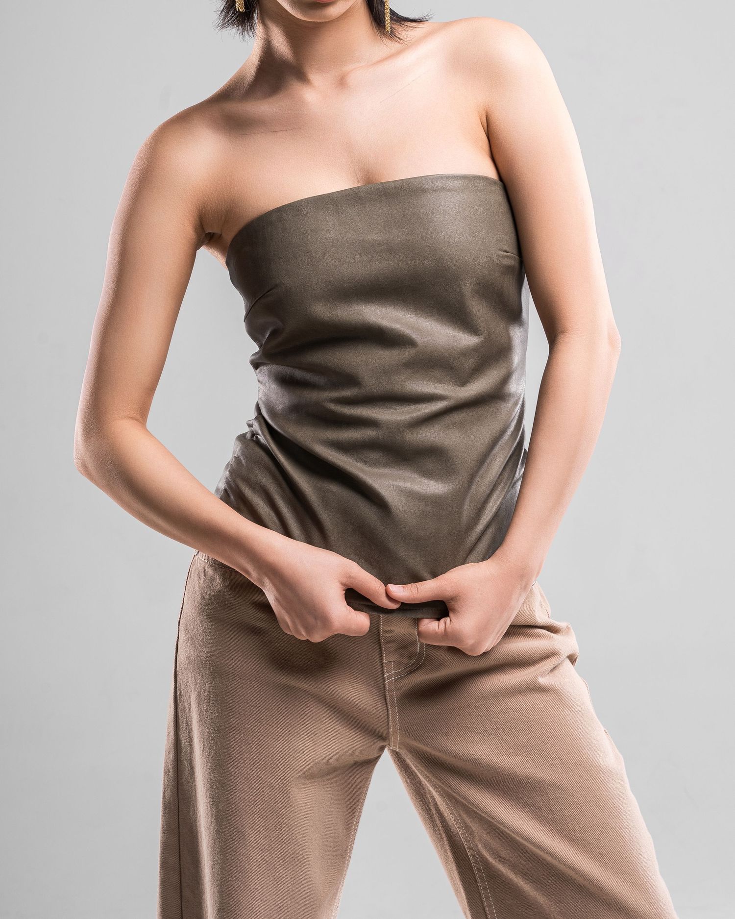 Fashion model poses in olive strapless top and beige pants against grey background.