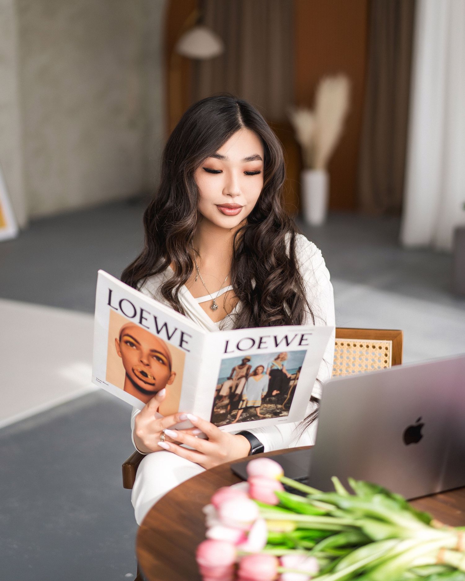 Person reading magazine at desk with laptop and tulips in modern interior setting.