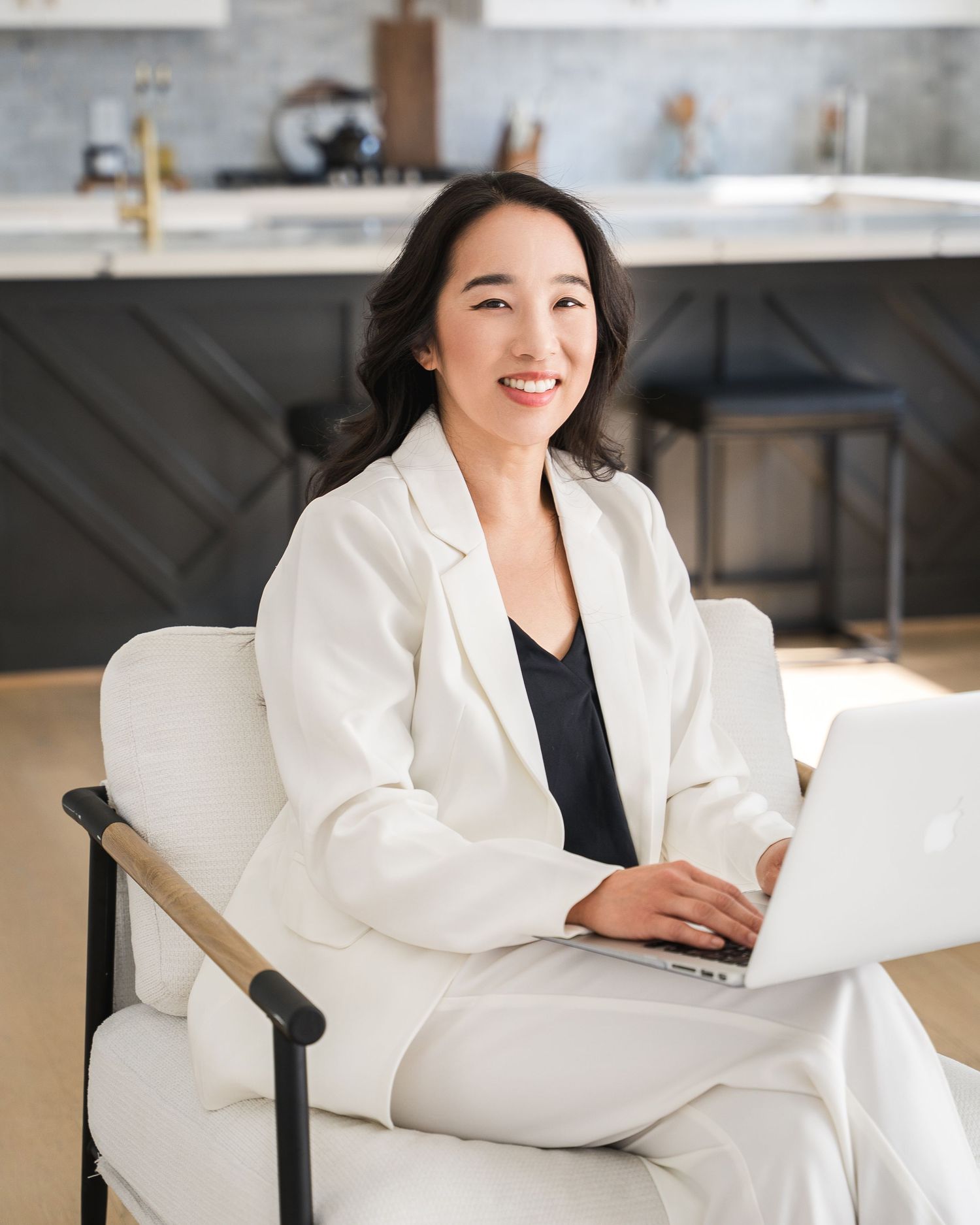 Professional working on laptop while seated in white chair in modern kitchen setting.
