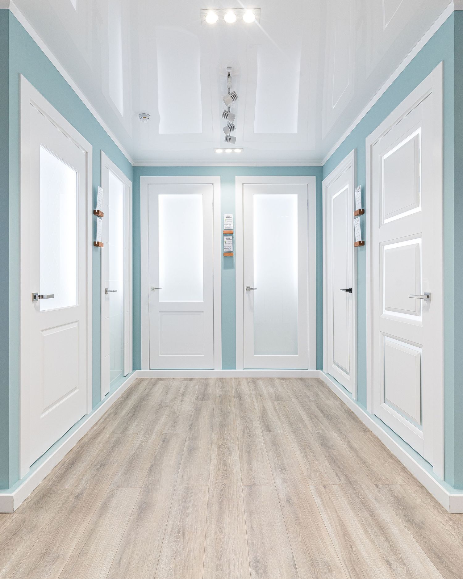 Clean modern corridor with light blue walls, white doors, and track lighting.