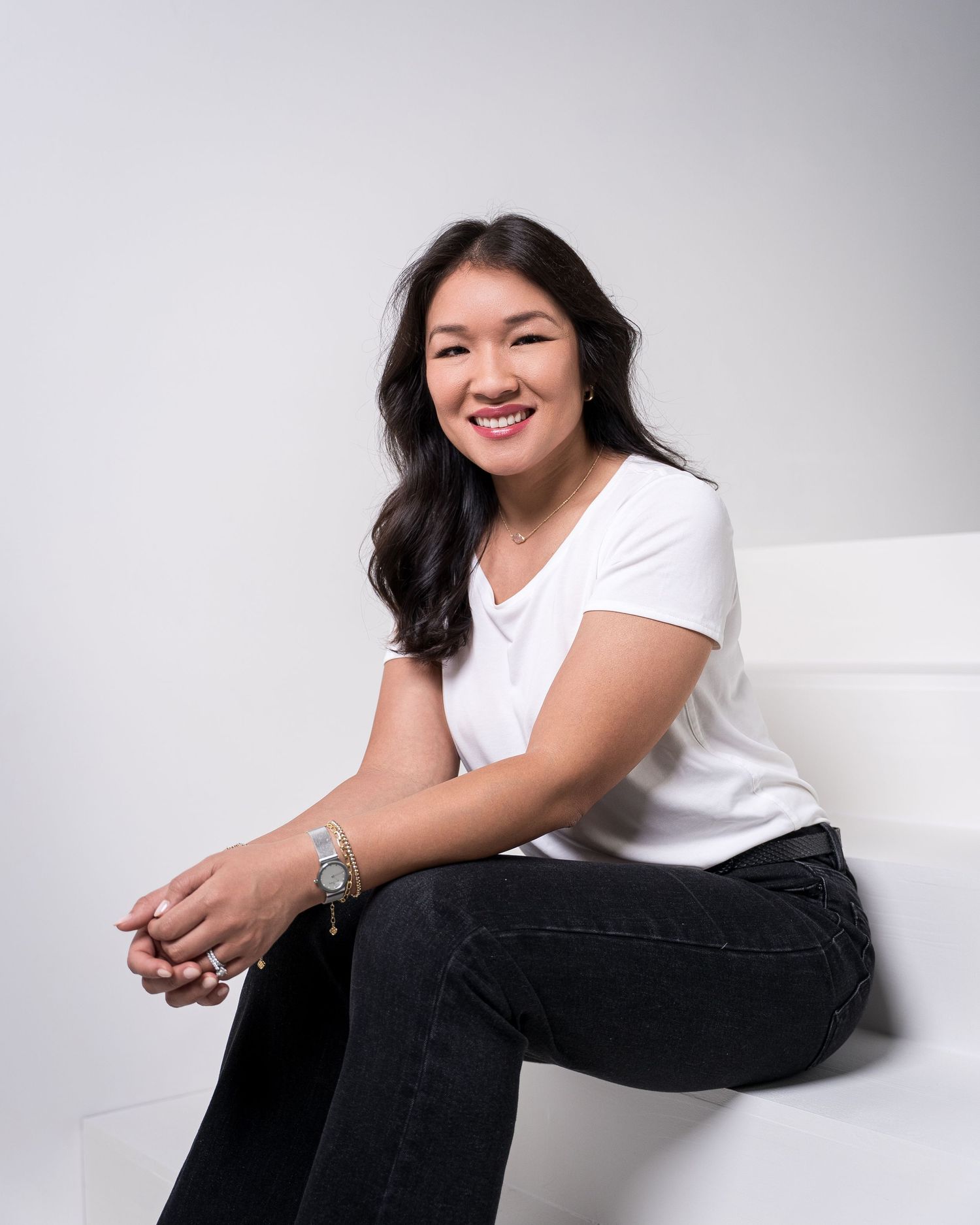 Woman in white top and dark pants sitting casually against white studio backdrop.