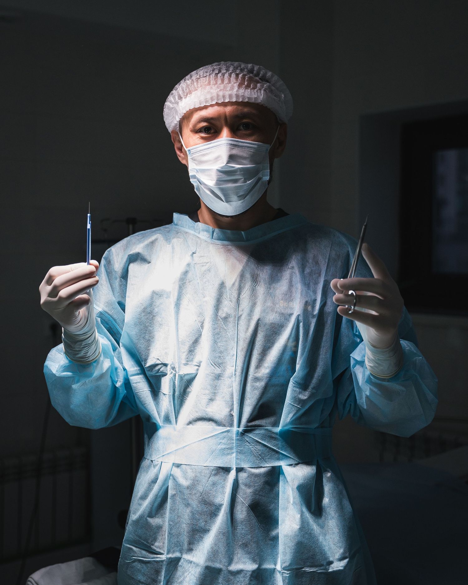 Medical professional in surgical gown and mask holding up surgical tools in operating room.