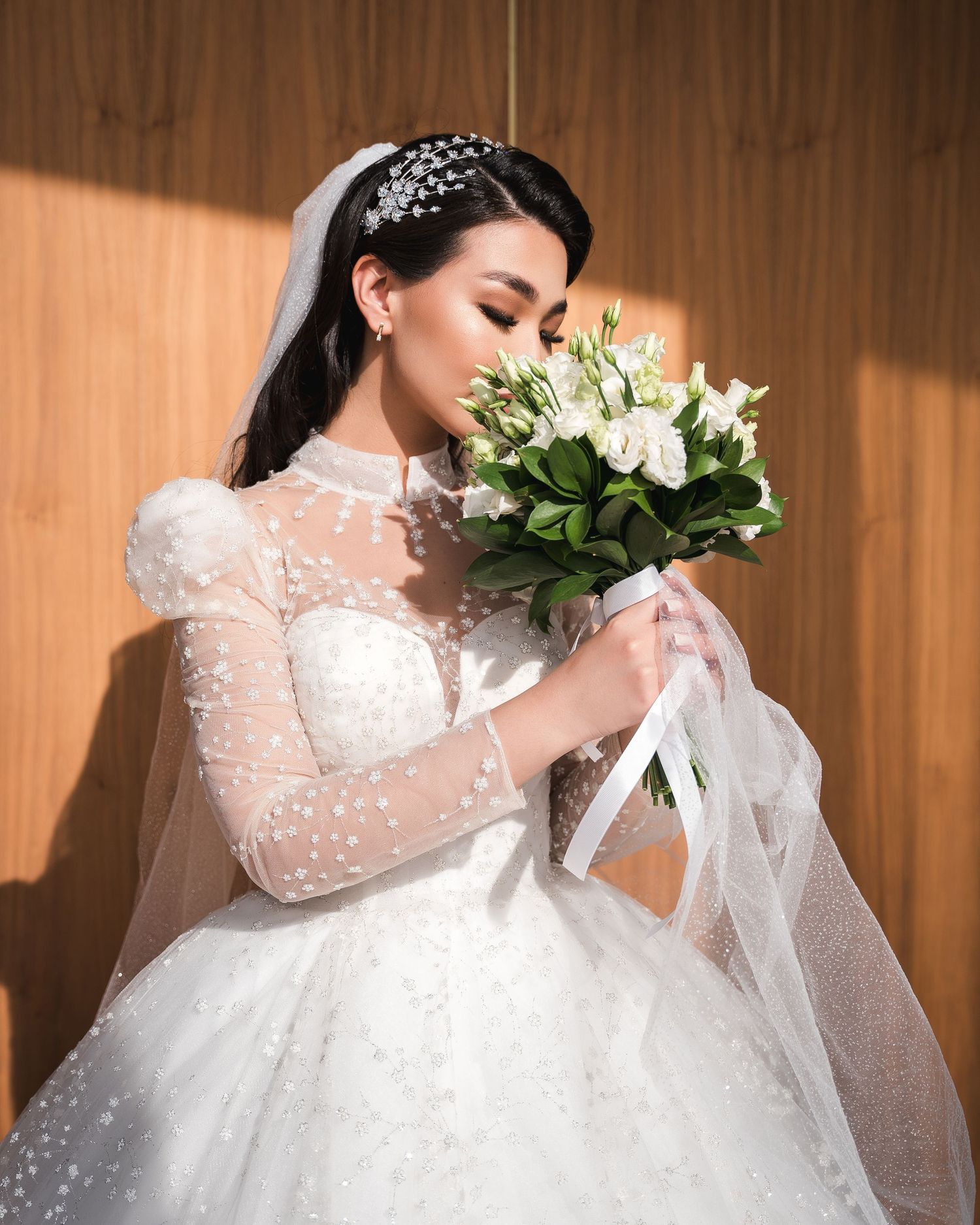 Elegant bride in pearl-embellished long-sleeve gown smells white flower bouquet with eyes closed.