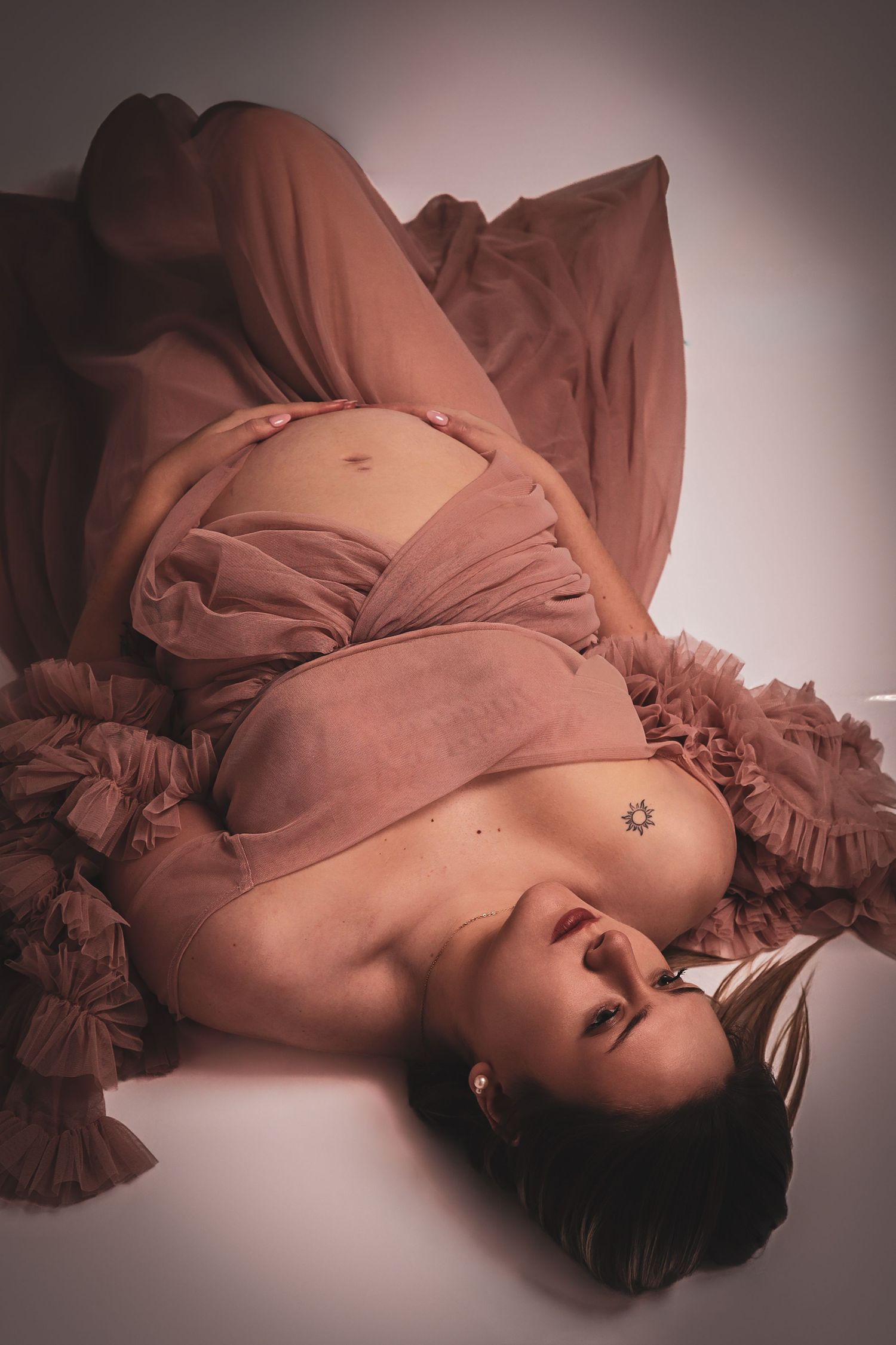Photographie de grossesse artistique en studio : femme enceinte allongée drapée dans un voile vaporeux rose, par Cindy Poulet Photographe