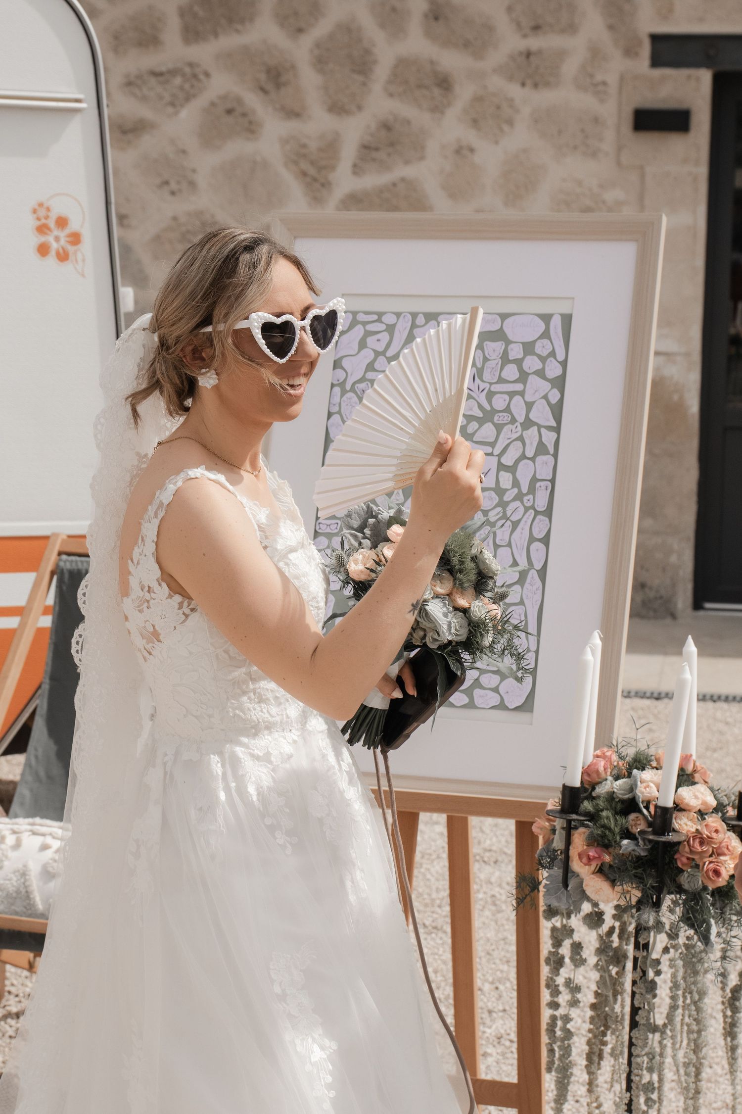 Mariée avec éventail et lunettes de soleil coeur devant tableau de mariage