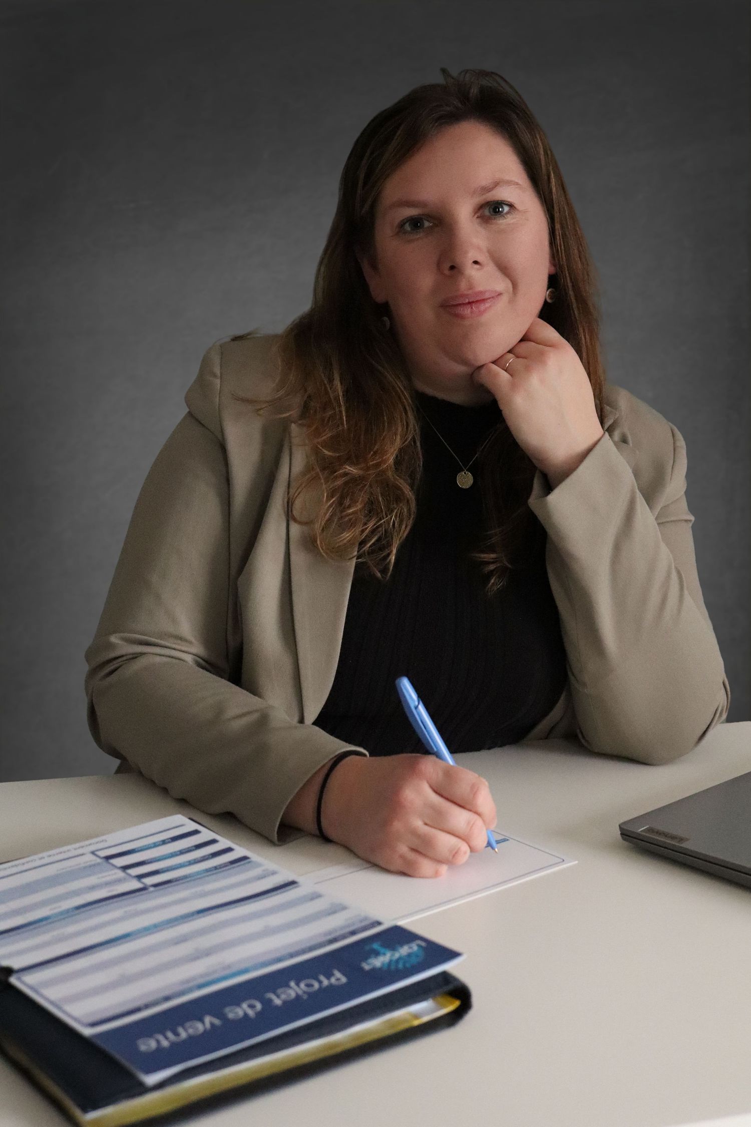 Portrait pro par Cindy Poulet Photographe : femme en blazer beige écrivant à son bureau avec ordinateur et dossier Projet de vente.