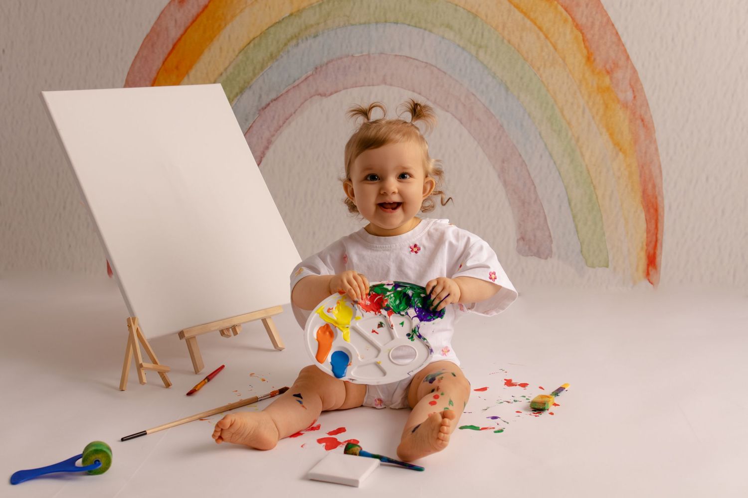 Bébé s'amusant avec de la peinture colorée devant un décor arc-en-ciel. Séance photo éveil artistique par Cindy Poulet à Septvaux, Aisne.