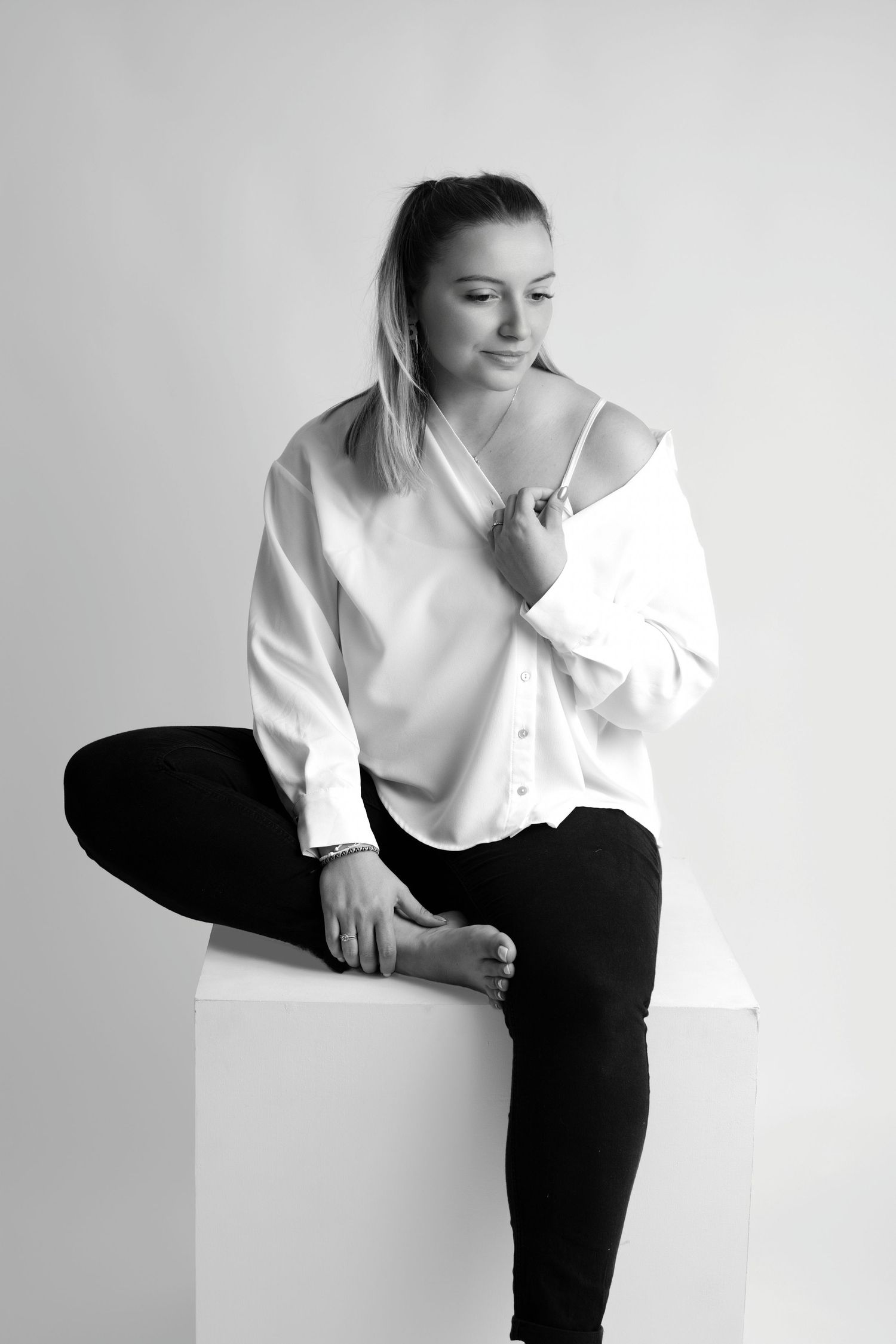 Portrait lifestyle en noir et blanc par Cindy Poulet Photographe : femme assise en studio sur un cube blanc, vêtue d'une chemise blanche et