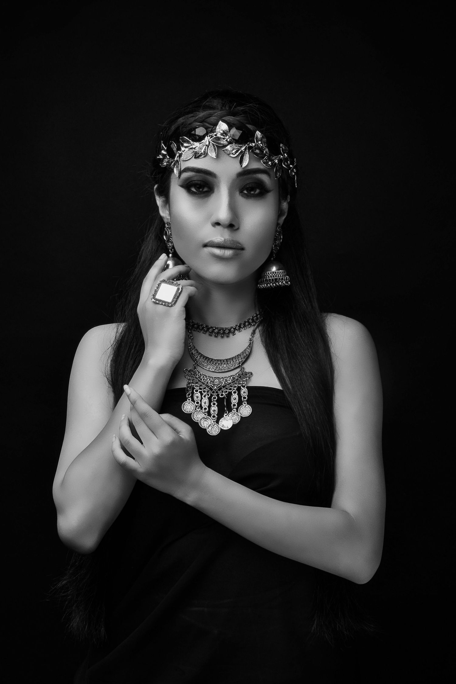 Black and white portrait of a woman in ornate jewelry, headpiece, and black dress against dark background.