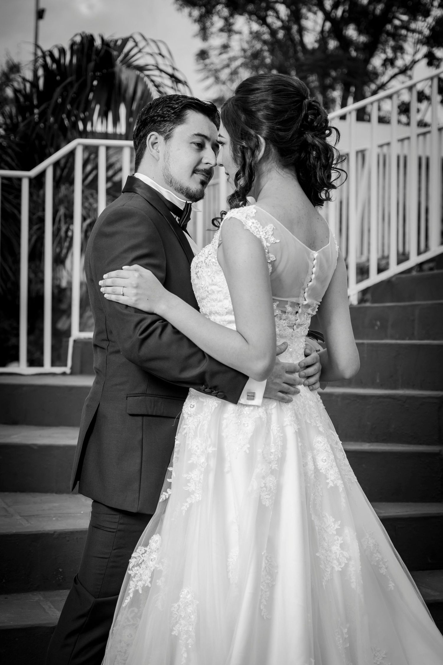 Black and white wedding photo of bride and groom embracing on outdoor steps, bride in lace gown, groom in dark suit.