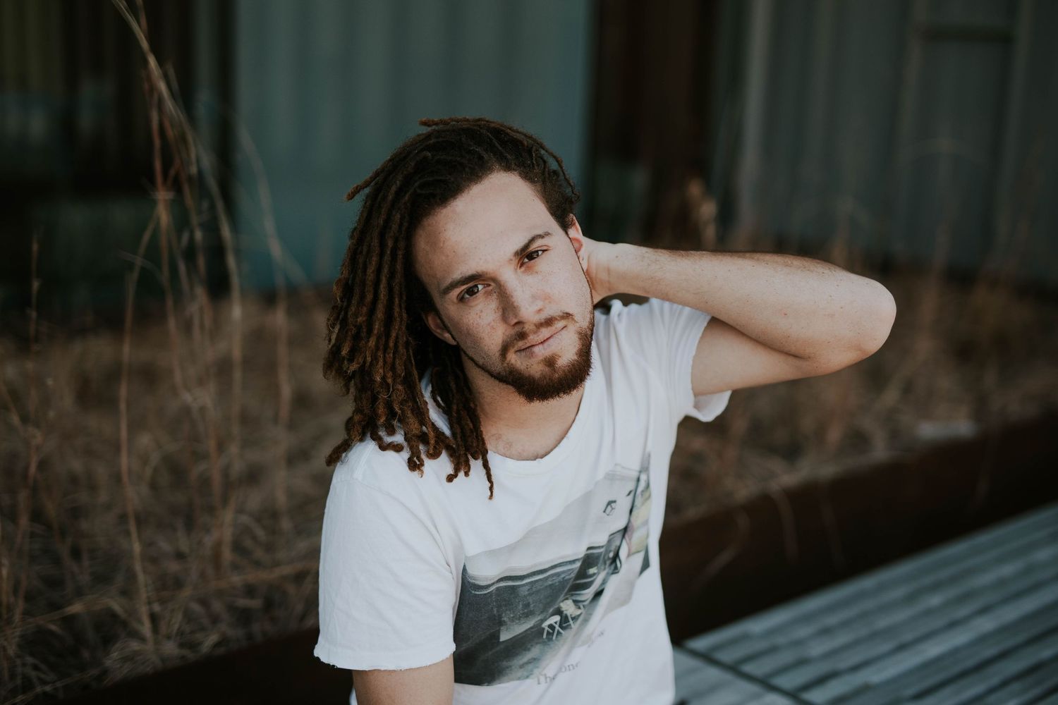 Young man with dreadlocks and beard wearing a white graphic t-shirt, posing outdoors in a relaxed portrait.