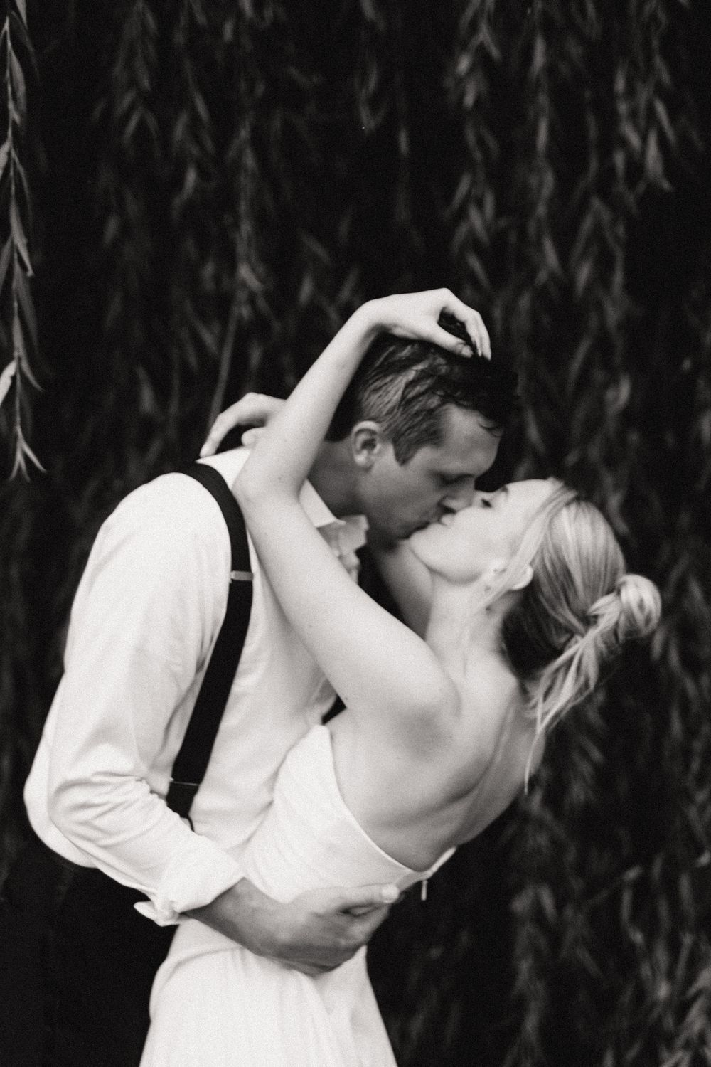 Black and white photo of a couple sharing a romantic kiss outdoors, man in suspenders embracing woman in white dress.