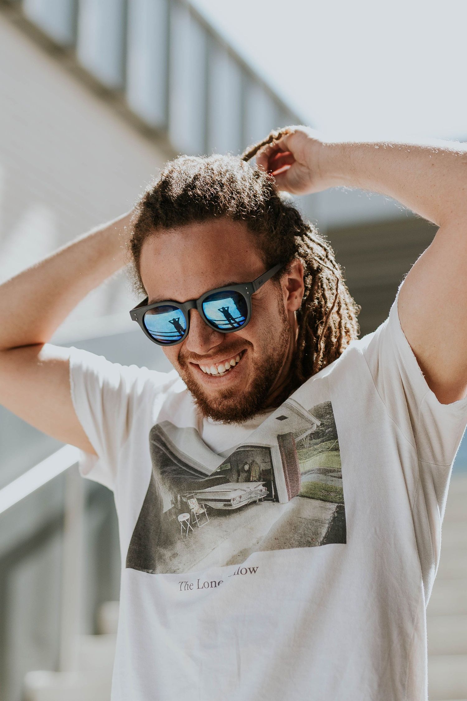 Smiling man with dreadlocks wearing blue mirrored sunglasses and a white graphic tee, hands behind his head.