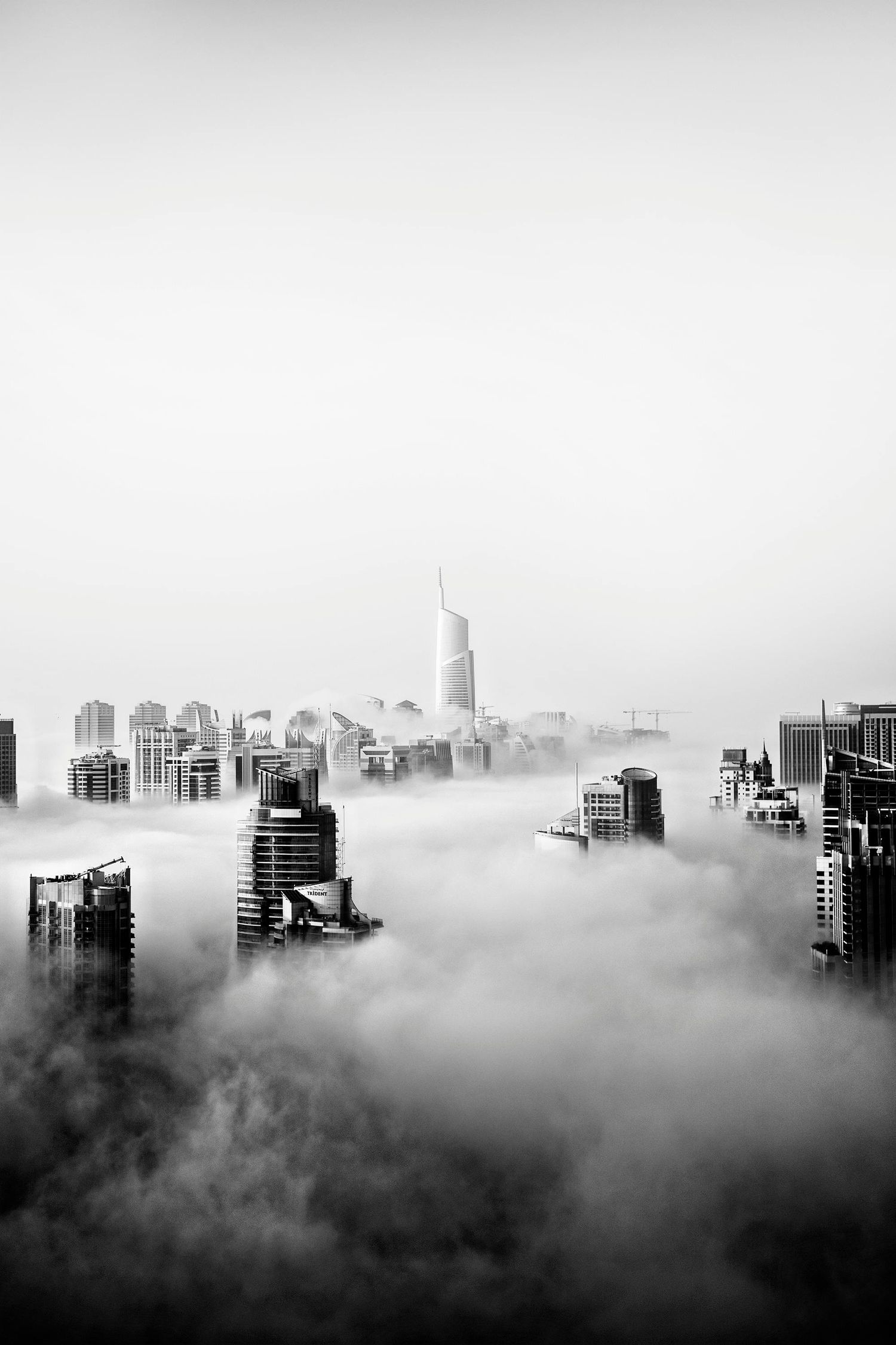 Black and white aerial view of city skyscrapers emerging through dense fog, creating a dramatic and mysterious urban landscape.