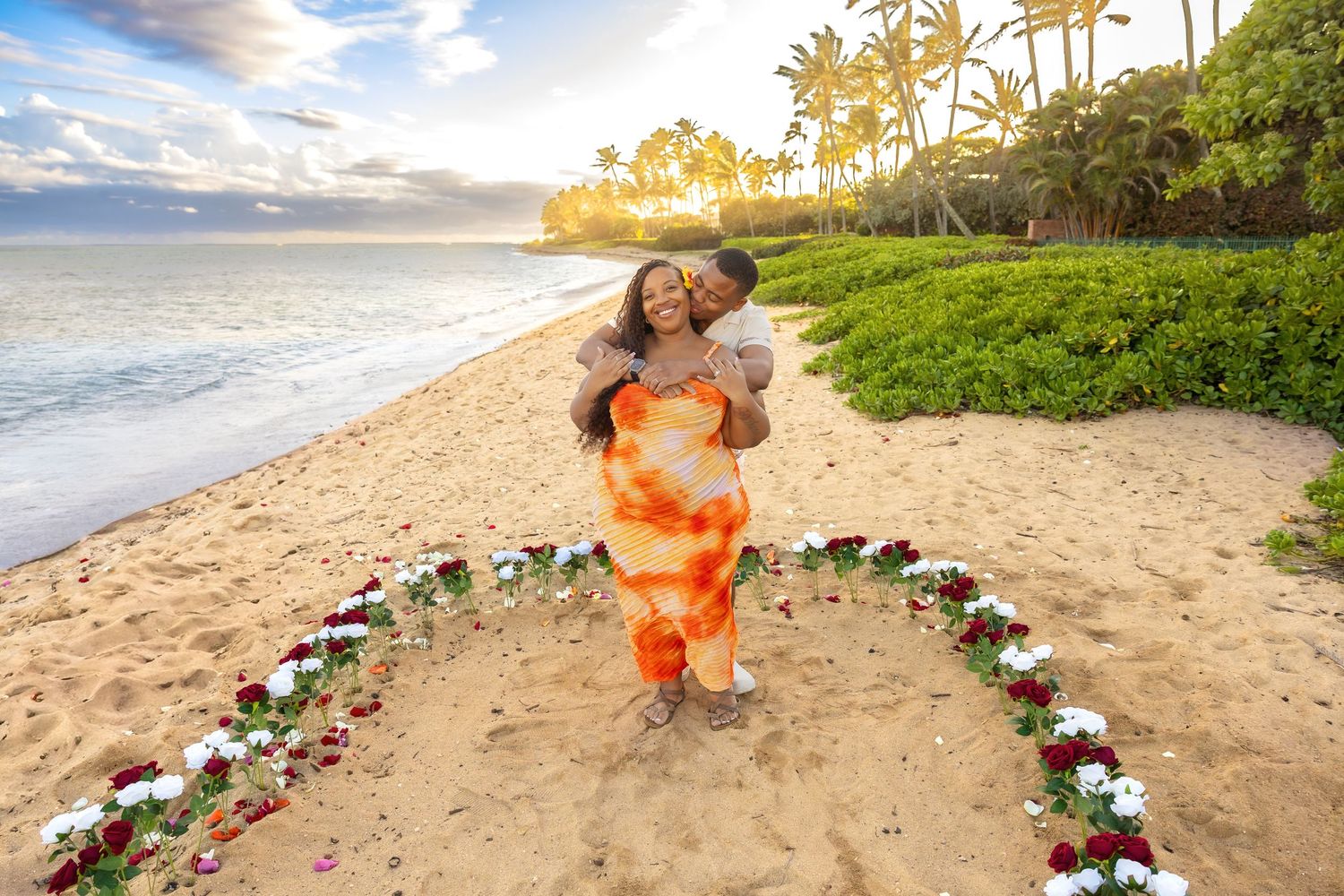 Happy couple embracing inside heart-shaped rose arrangement on Hawaiian beach at sunset with palm trees.
