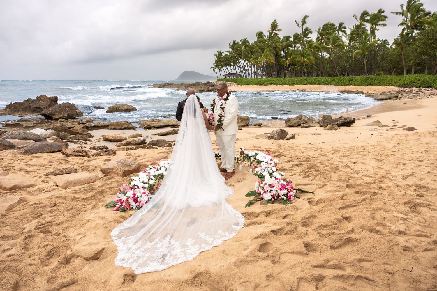 Bride and groom exchange vows on sandy Hawaiian beach with floral arrangements and palm trees.