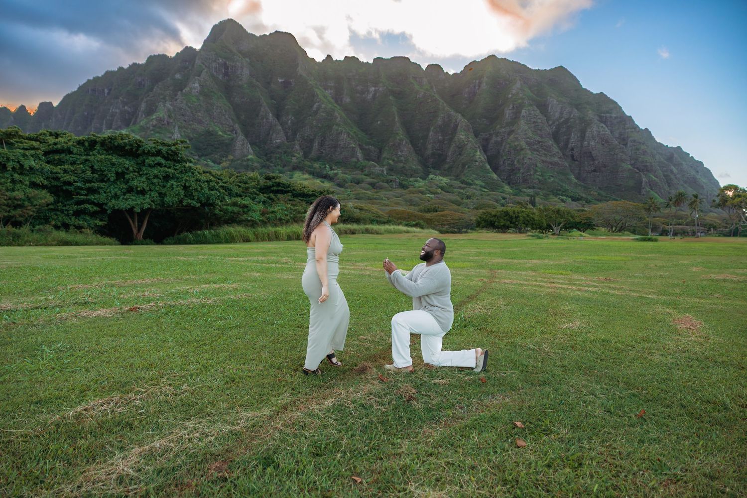 Man proposing on one knee to woman in white dress with dramatic mountain backdrop in Hawaii.