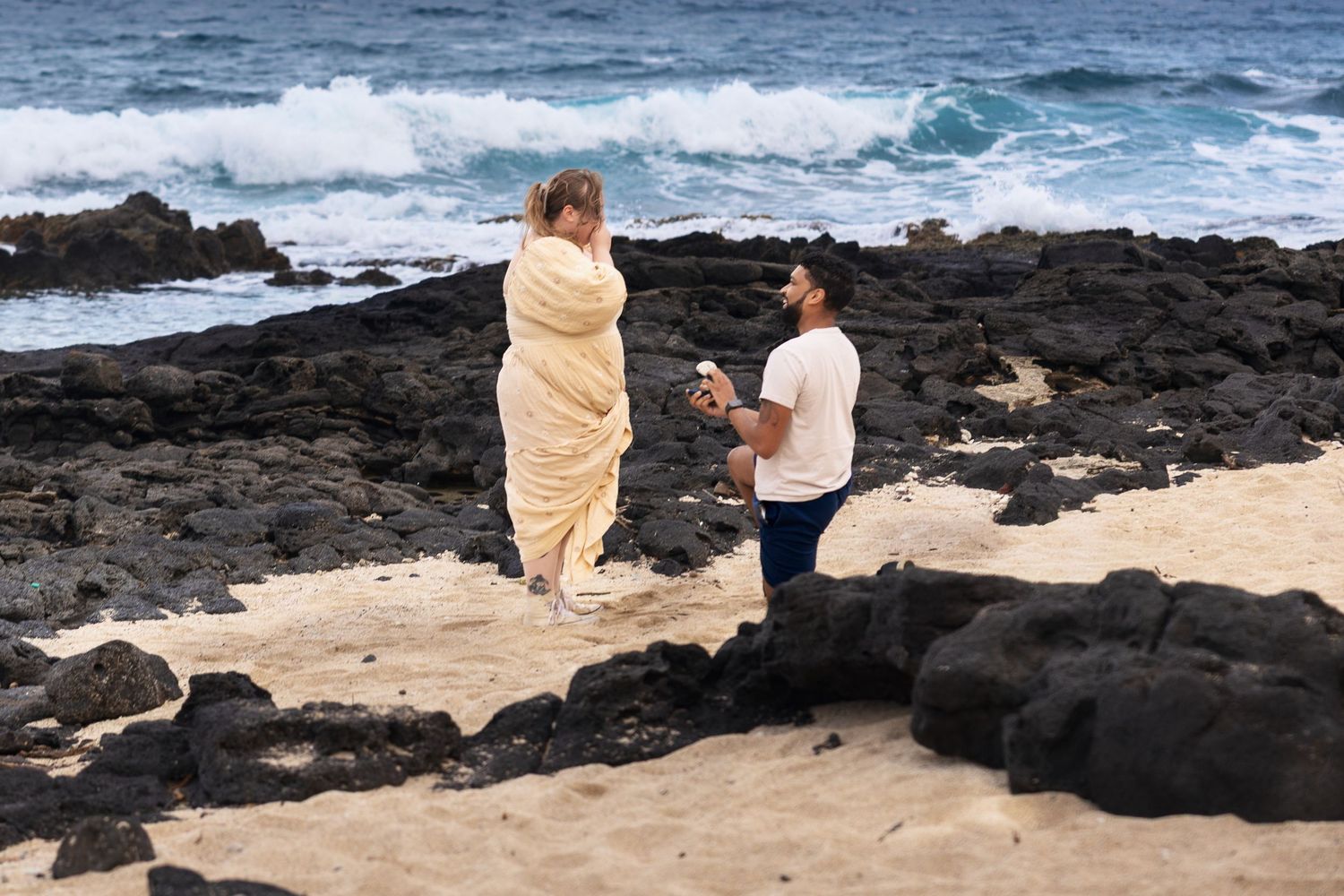 Romantic beach proposal with man on one knee holding ring box for woman wrapped in blanket by rocky shore.