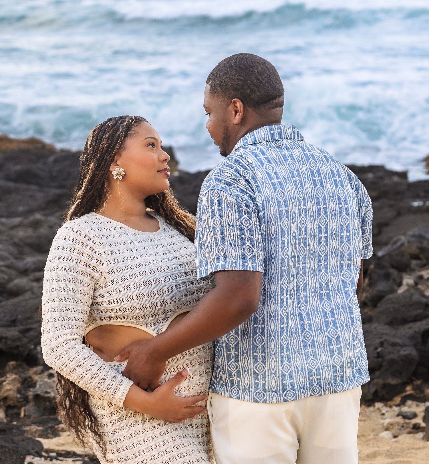 Couple sharing tender moment by rocky Hawaiian shoreline with ocean waves in background.