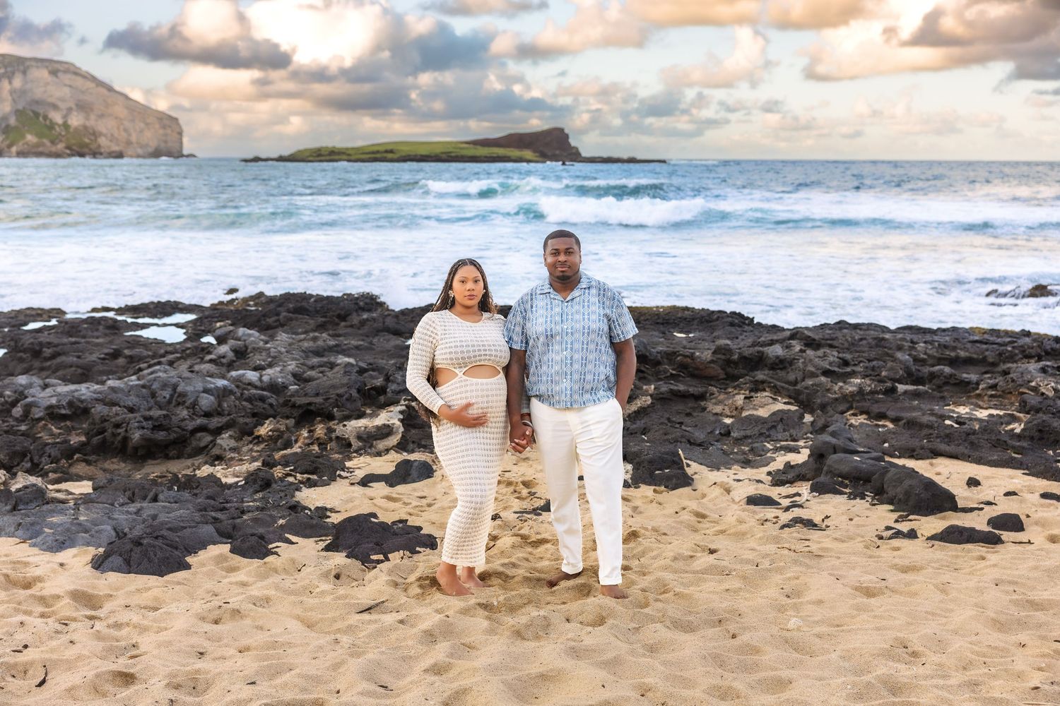 Couple poses on sandy Hawaiian beach at sunset with rocky shoreline and island in background during maternity shoot.