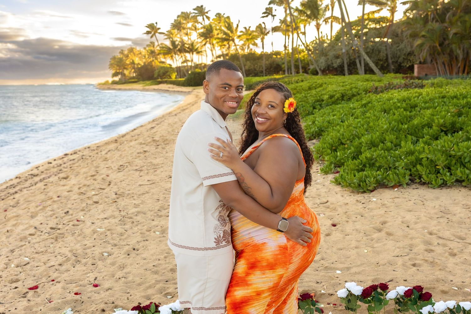 Joyful couple posing together on Hawaiian beach at golden sunset, woman in orange tie-dye dress with flower in hair.