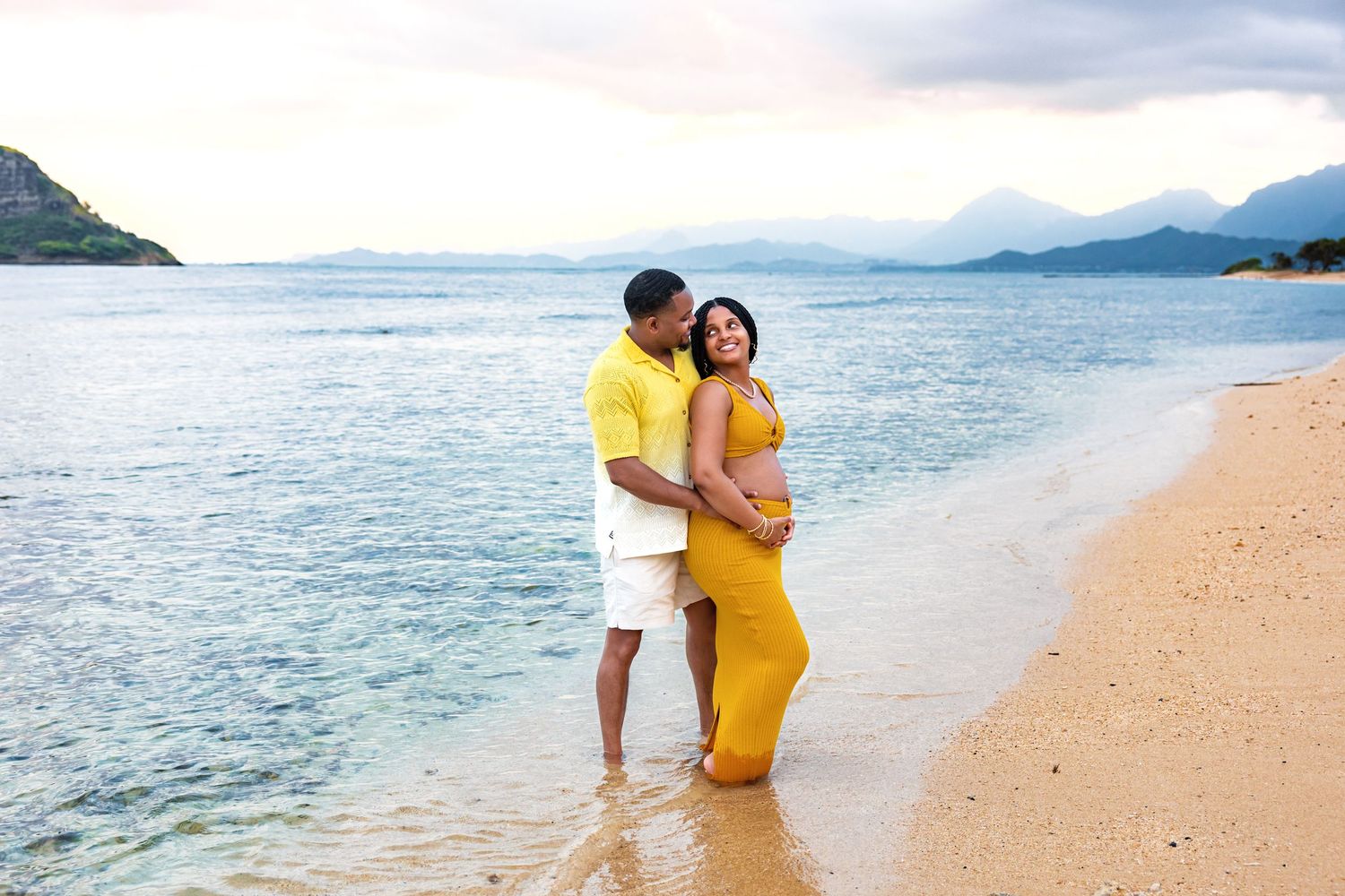 Couple embraces on shallow Hawaiian shoreline at dusk, man kisses partners head as she cradles her baby bump.