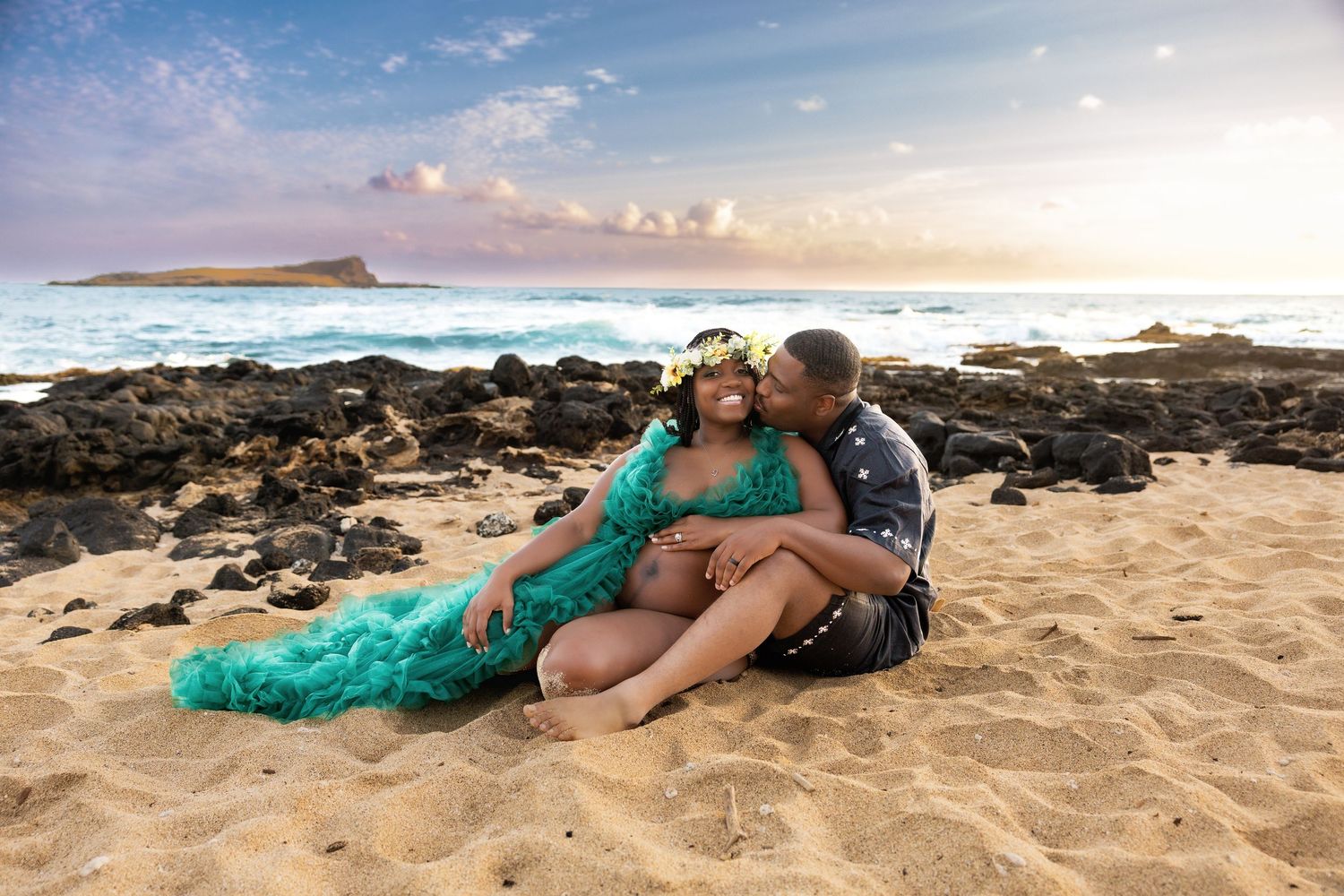 Couple sharing a kiss while sitting on sandy Hawaiian beach at sunset in teal maternity gown.