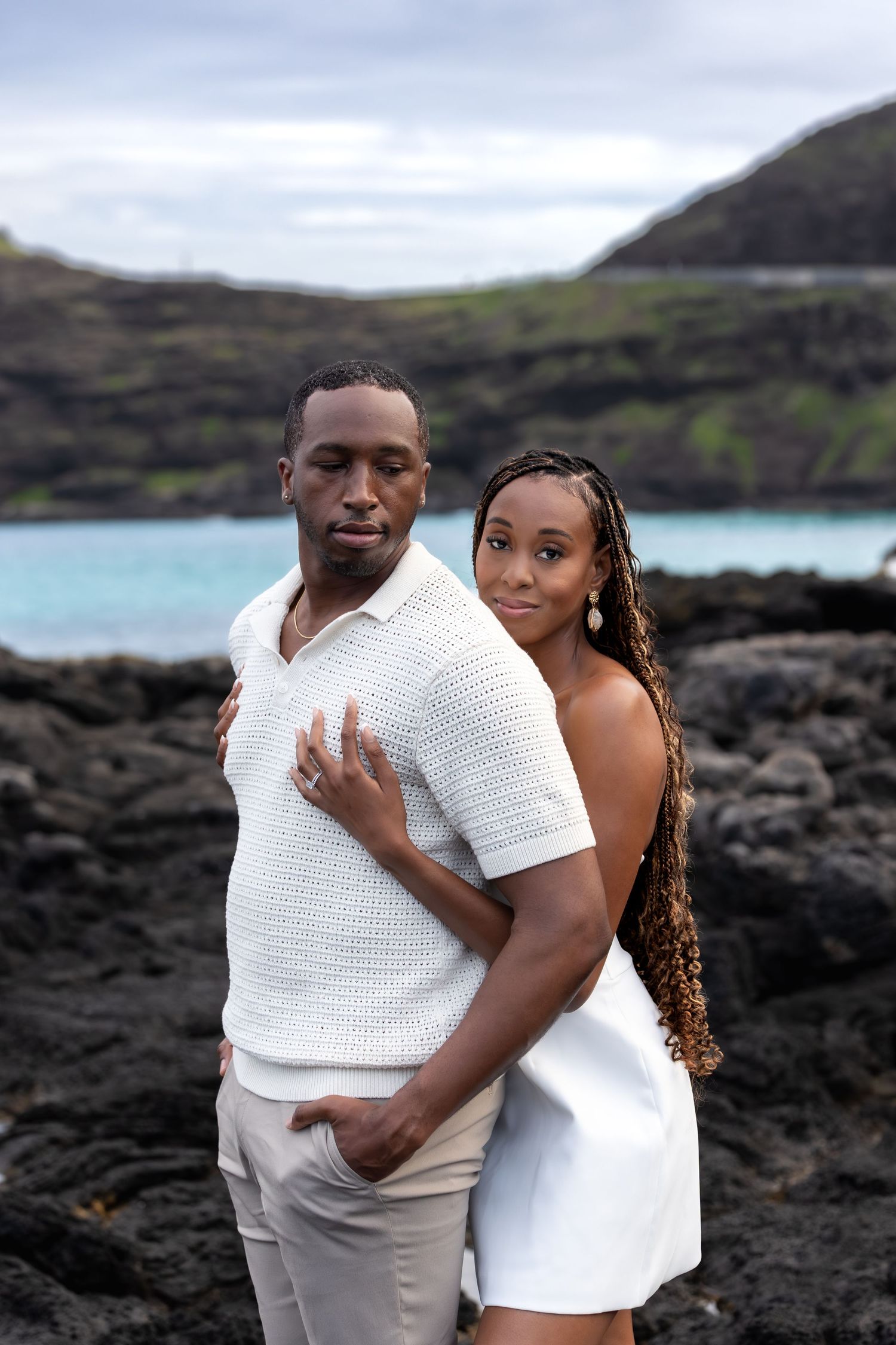 Couple in white outfits posing on black lava rocks with blue ocean behind them.