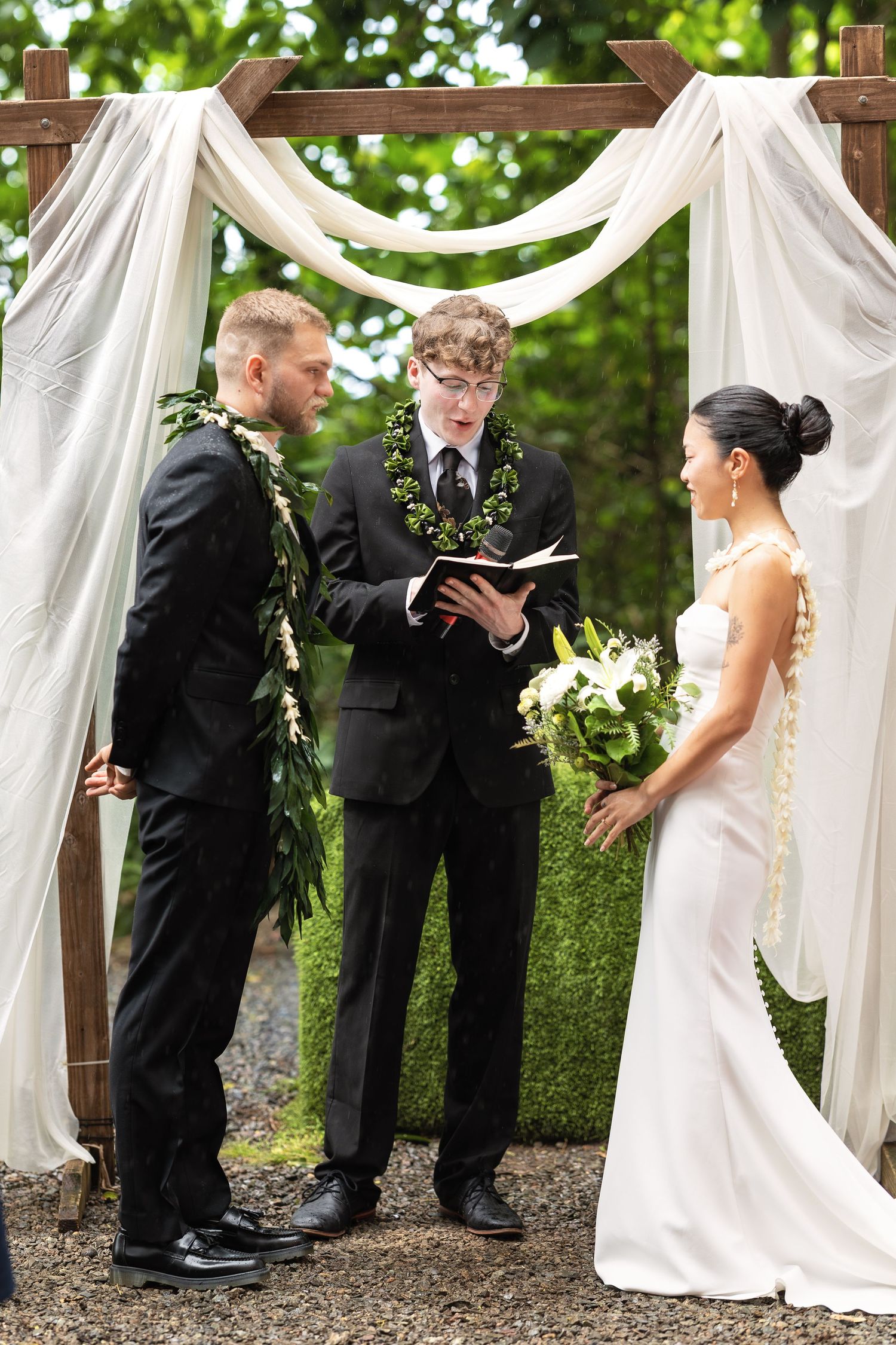 Bride and groom listening to officiant read vows under a white draped wooden arch at an outdoor wedding.