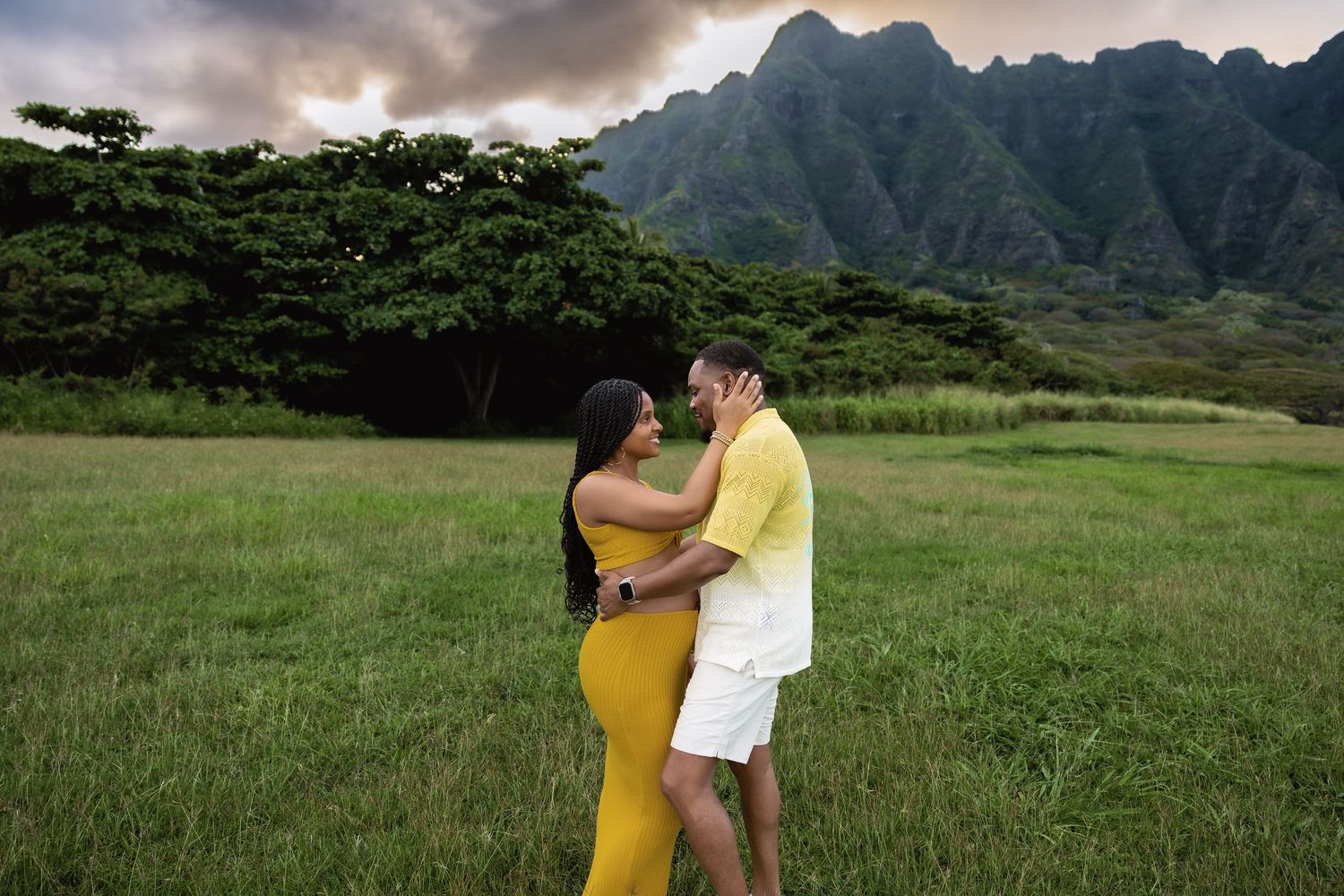 Expecting couple shares an intimate moment in grassy Hawaiian field with dramatic mountain range at golden hour.