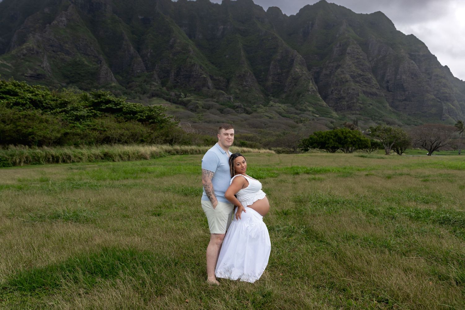 Couple posing together in lush green field with stunning Ko'olau mountain range in background.