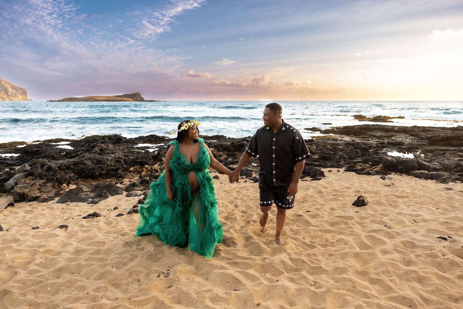 Couple holding hands on sandy Hawaiian beach at sunset wearing elegant teal maternity gown.