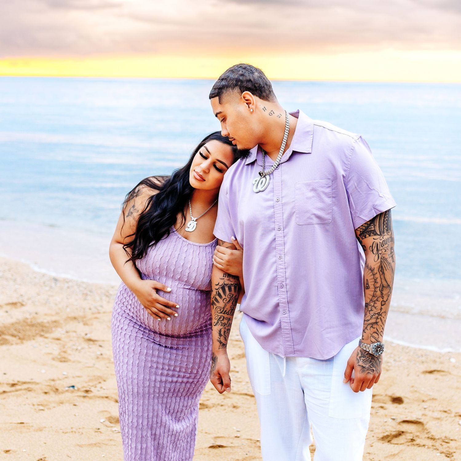 Expecting couple in matching lavender outfits embracing on a sandy beach at colorful sunset.
