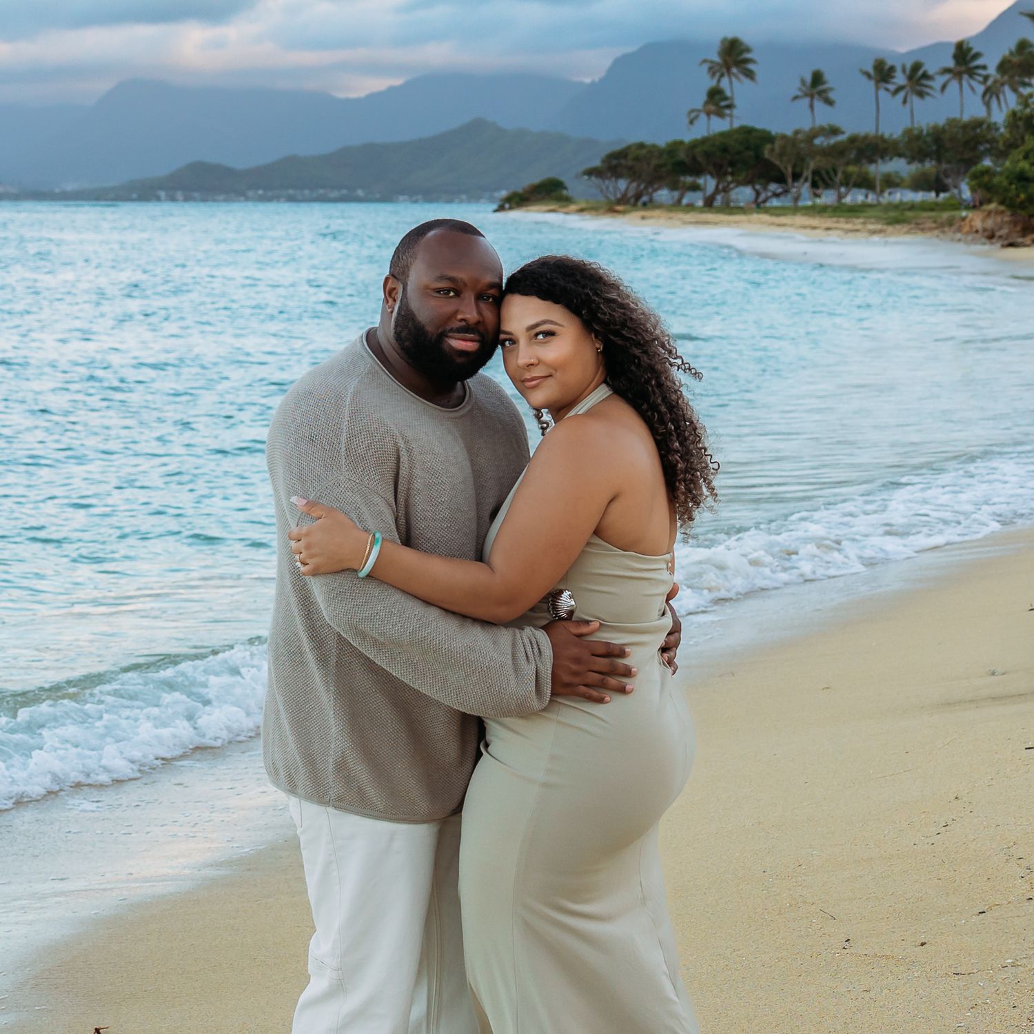 Smiling engaged couple posing together on sandy Hawaiian beach at golden sunset hour.