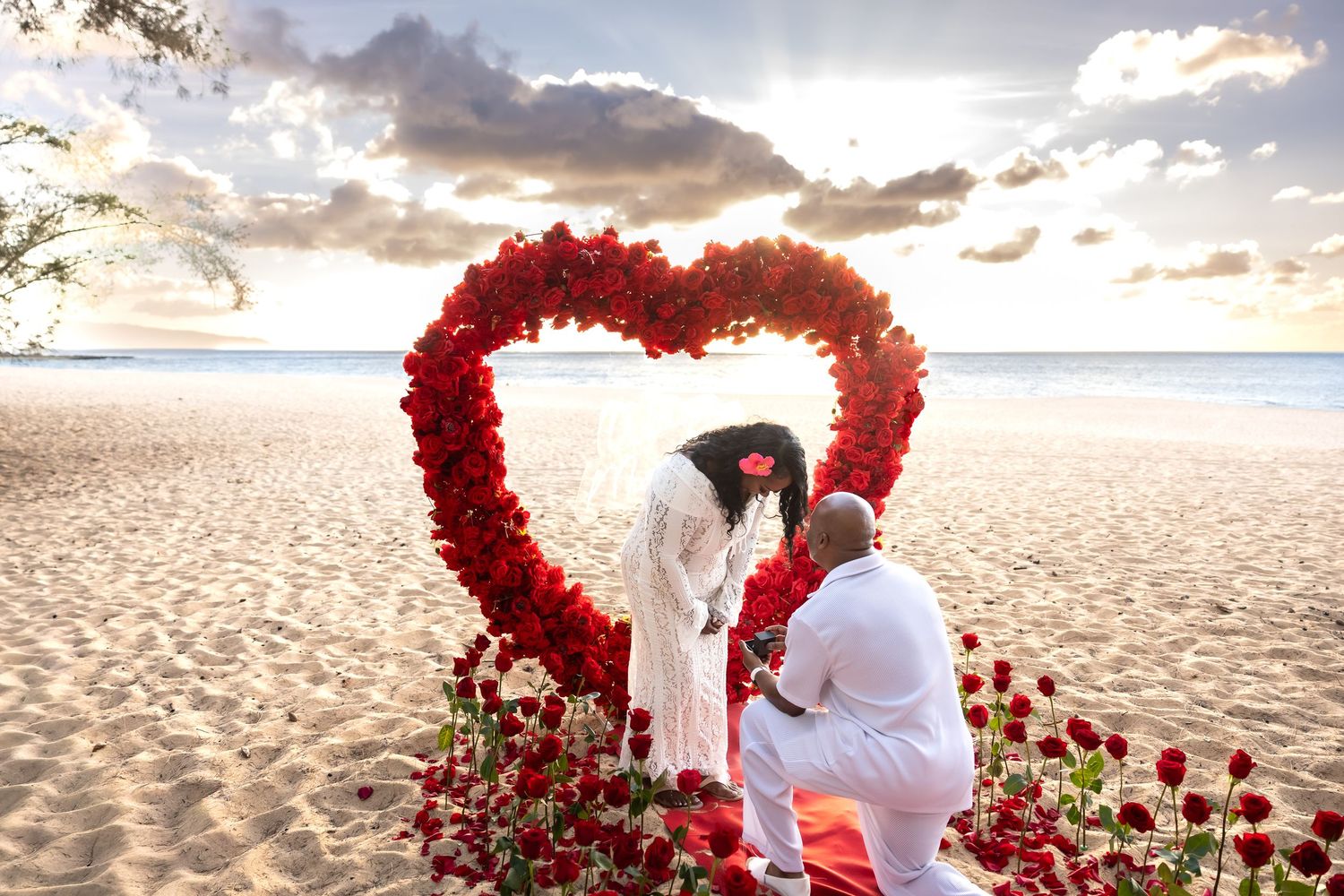Couple sharing magical marriage proposal moment inside red rose heart arch on sandy beach at sunset.