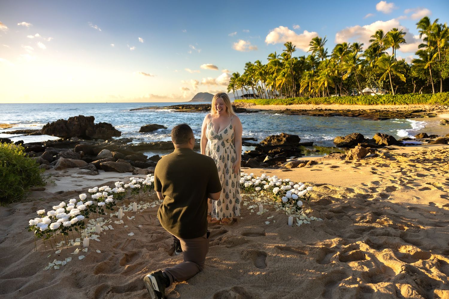 Man on one knee proposing to woman on tropical beach surrounded by white floral arrangement at sunset.