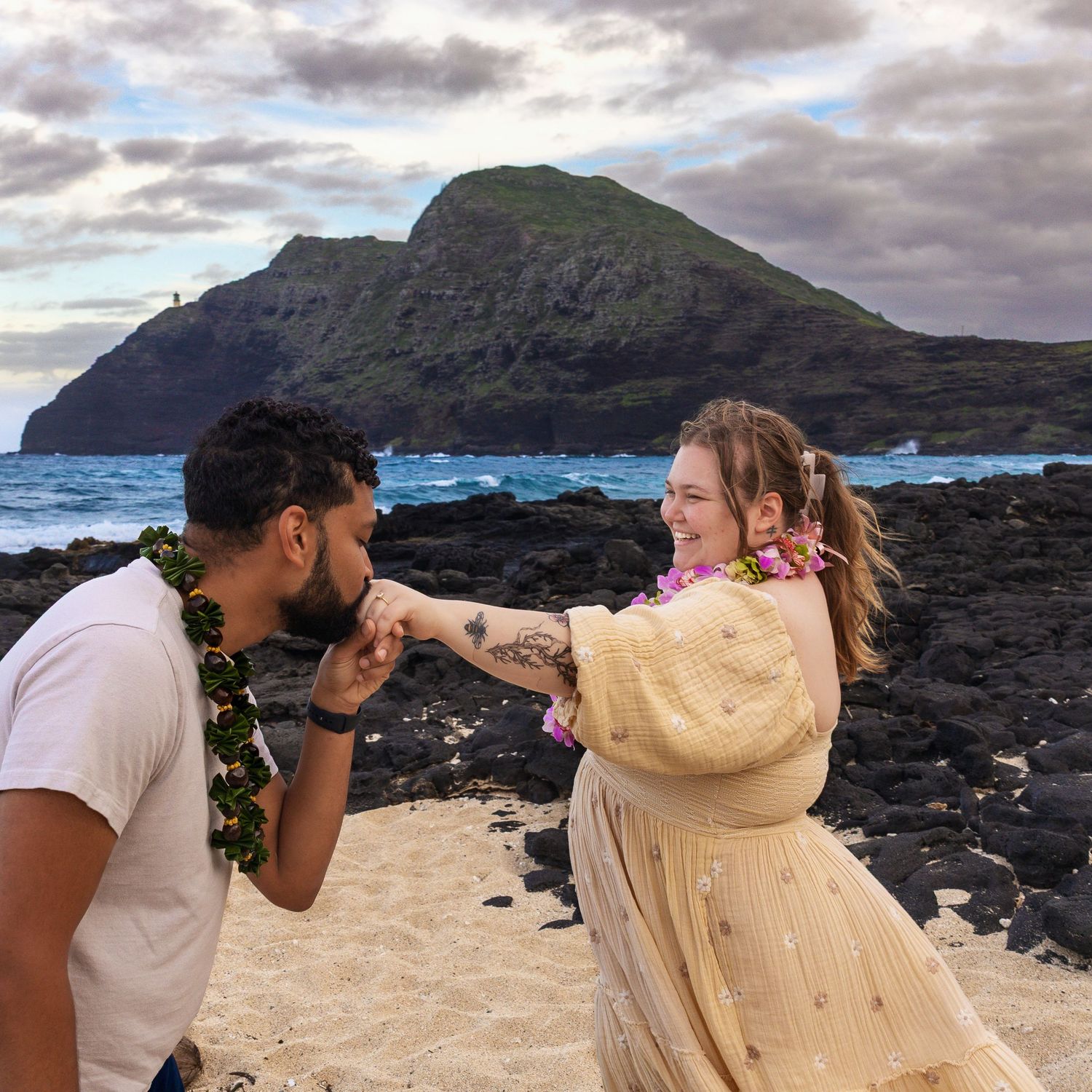 Newly engaged couple celebrating on Hawaiian beach, man kissing fiancee's hand with Makapuu island backdrop.