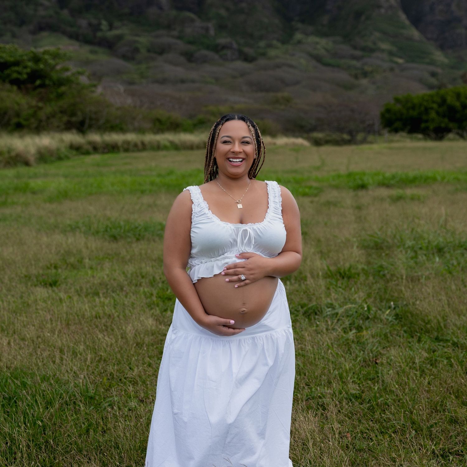 Smiling pregnant woman in white crop top and skirt holding baby bump in Hawaii field.