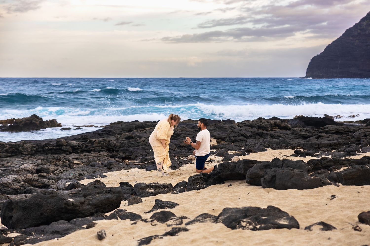 Man kneeling in proposal on rocky Hawaiian beach with ocean waves and dramatic cliffs in background.