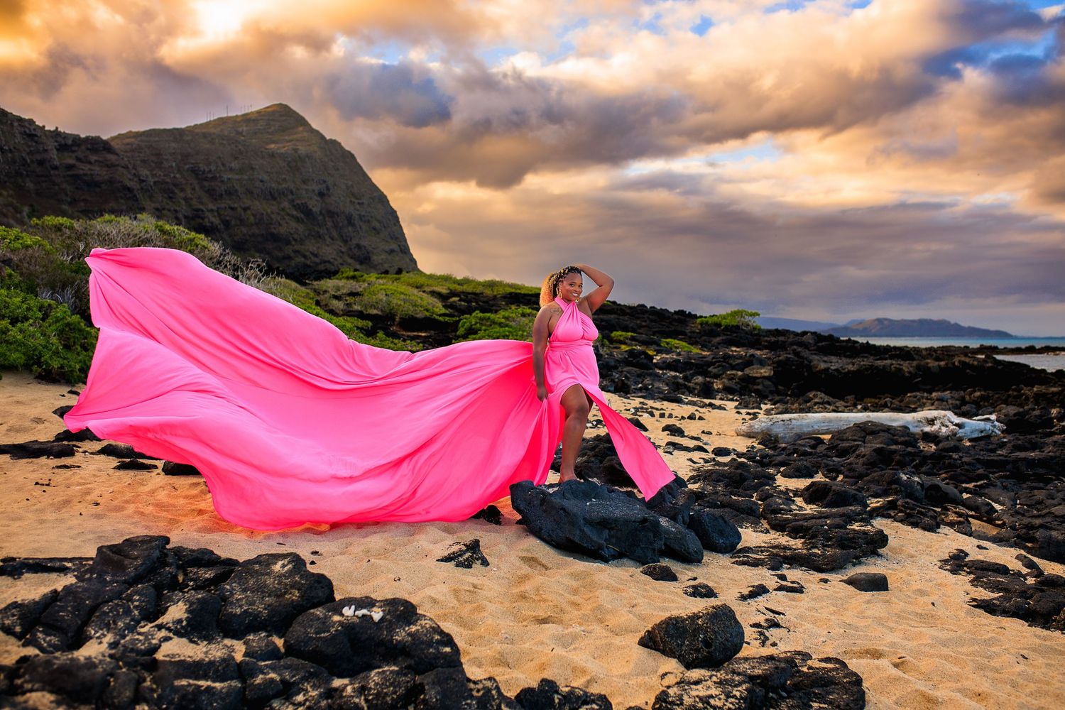 Woman in flowing pink gown posing dramatically on a rocky Hawaiian beach at golden sunset.