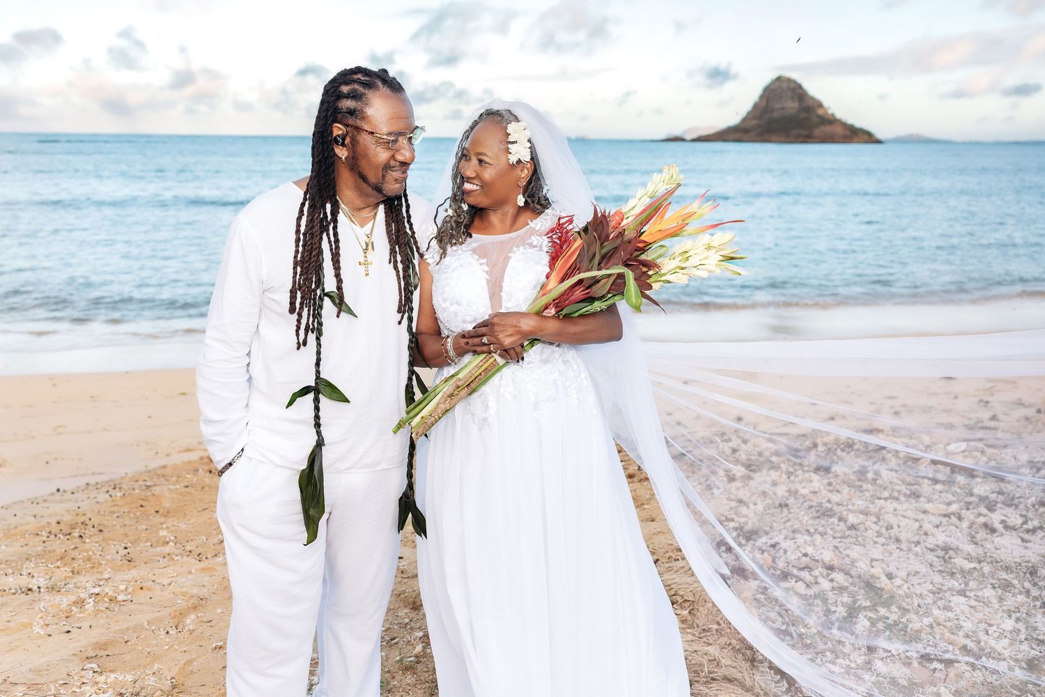 A couple in white attire poses on a Hawaiian beach with Chinaman's Hat island in the background.