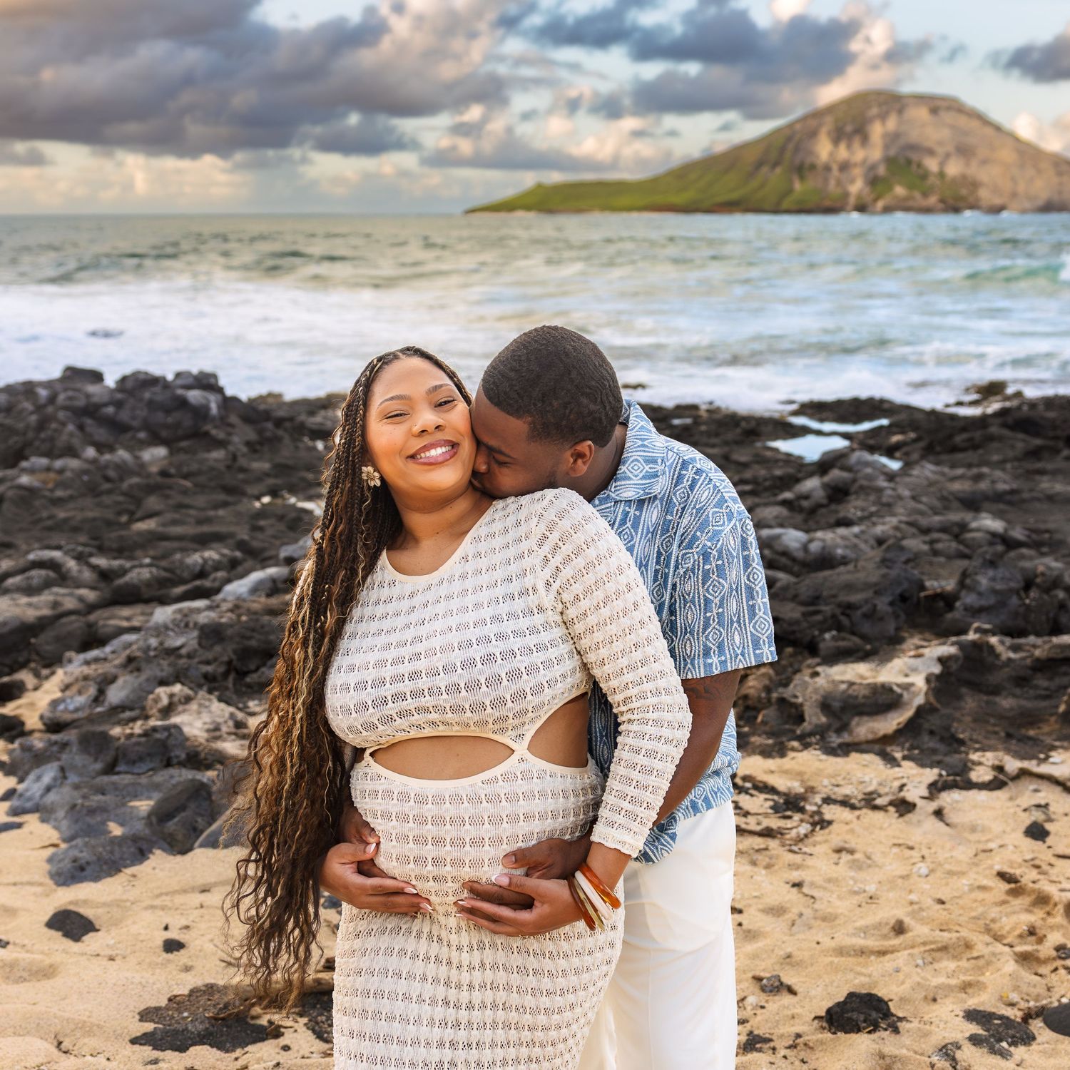 Couple embracing on Hawaiian beach at sunset with rocky shoreline and island in background.