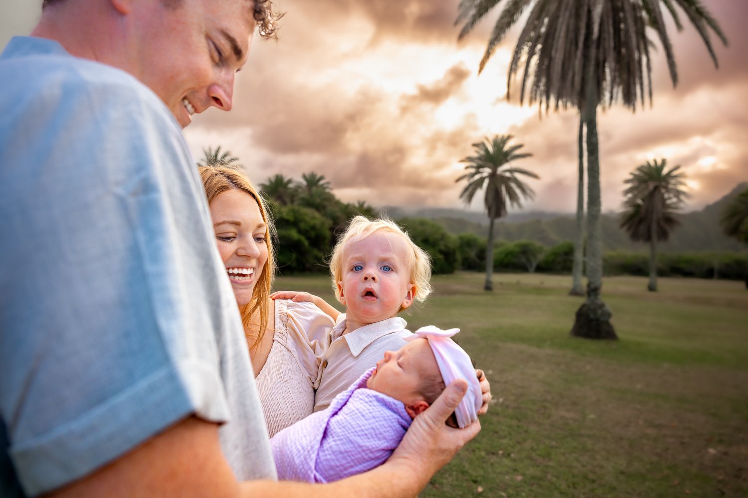 Young family with toddler and newborn baby smiling outdoors with palm trees and dramatic sky.