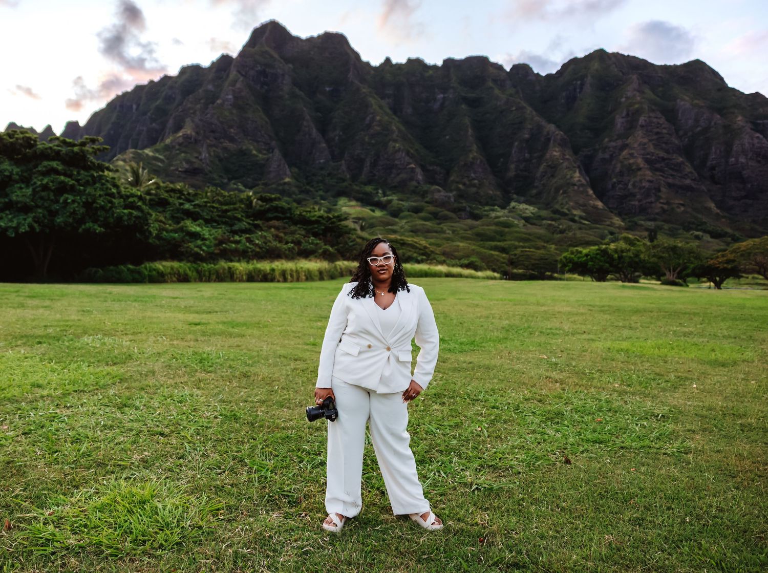 Woman in white pantsuit holds camera while standing in Hawaiian field with jagged mountain range at dusk.