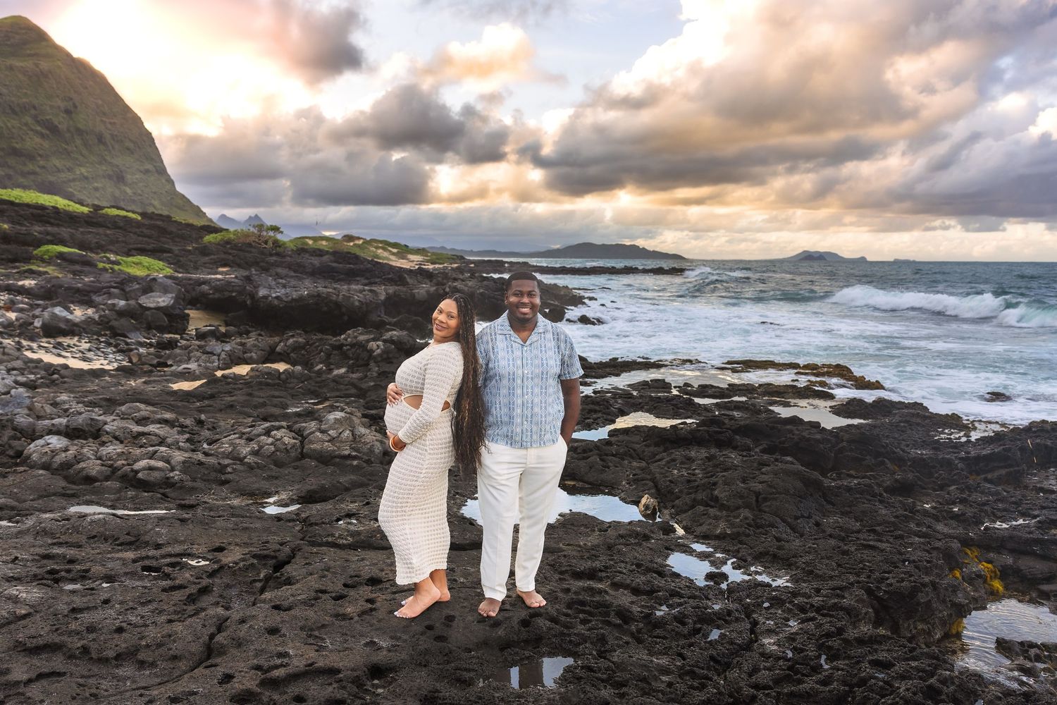 Pregnant woman in white dress and partner stand on volcanic rocks at Hawaiian coastline during dramatic sunset.